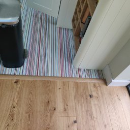 Drewno - Jasny parkiet z widocznymi słojami drewna, przechodzący w kolorową, pionowo pasiakową podłogę w pomieszczeniu z otwartymi drzwiami i czarnym koszem na śmieci.