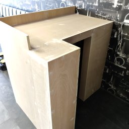 Drewno - Surowa konstrukcja z jasnej sklejki, prawdopodobnie fragment baru lub lady, widoczna na tle ściany z fototapetą przedstawiającą panoramę Londynu; widoczne ślady szpachli i niedoskonałości wykończenia.