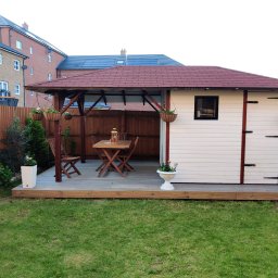 Drewno - Zadaszona drewniana konstrukcja z altaną, tarasem i domkiem narzędziowym, wyposażona w drewniany stół i krzesła, otoczona zielonym trawnikiem i drewnianym ogrodzeniem.