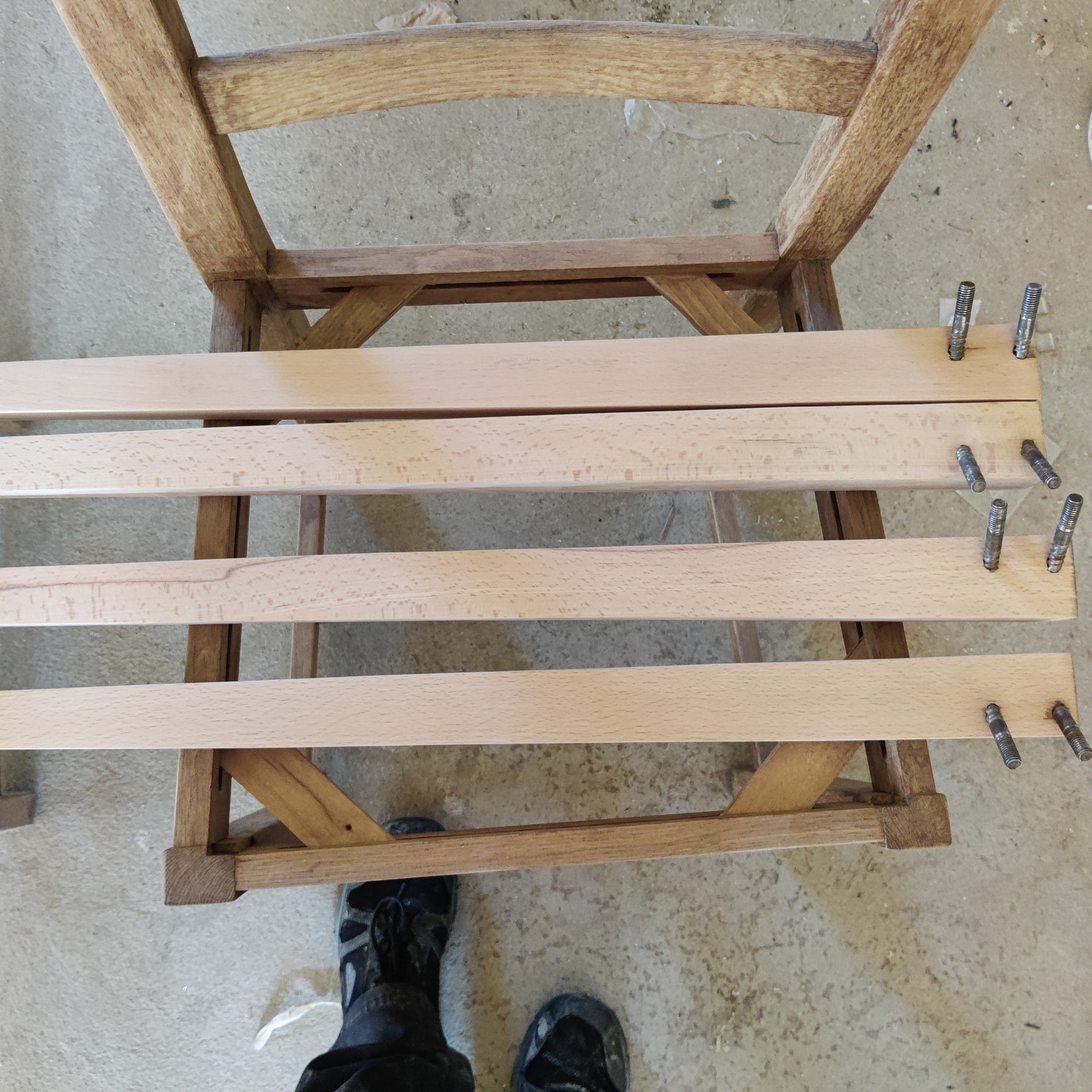 Surowe, jasne listwy drewniane z metalowymi kołkami ułożone na częściowo odnowionym, brązowym stelażu drewnianym, widoczne buty robocze w tle.