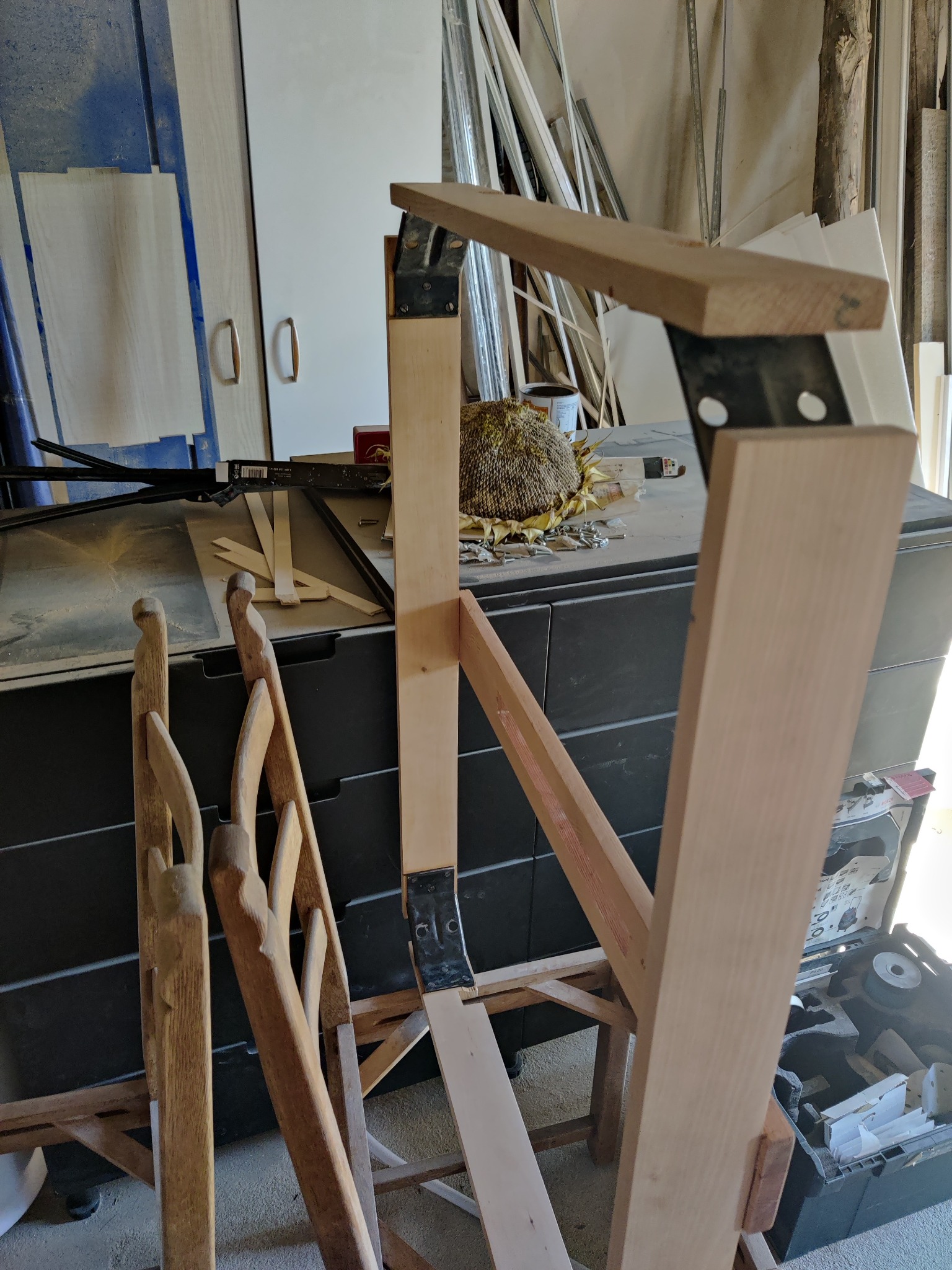 Fragment konstrukcji z surowego drewna łączonego metalowymi kątownikami, obok leżą rzeźbione elementy drewniane, w tle widoczny zasuszony słonecznik i narzędzia.