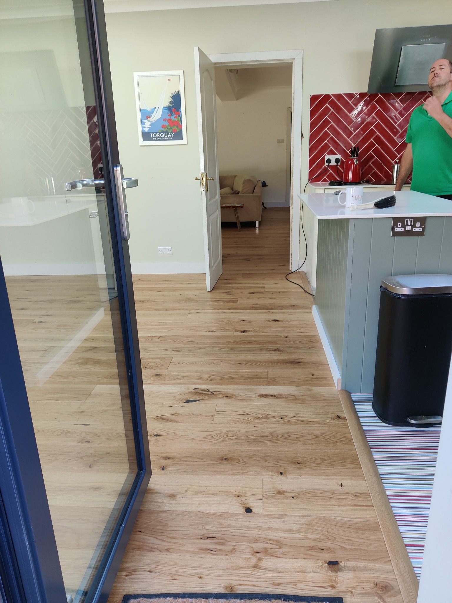 Jasne panele podłogowe w nowocześnie urządzonej kuchni, widok z perspektywy otwartego okna balkonowego na wnętrze z wyspą kuchenną i czerwonymi płytkami na ścianie.