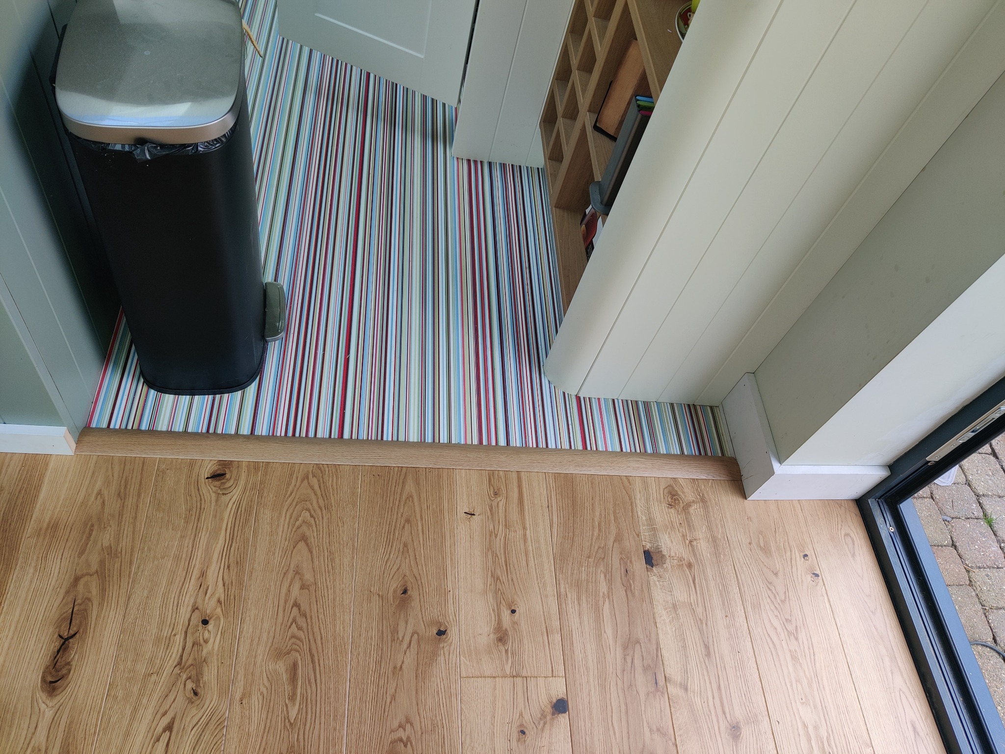 Jasny parkiet z widocznymi słojami drewna, przechodzący w kolorową, pionowo pasiakową podłogę w pomieszczeniu z otwartymi drzwiami i czarnym koszem na śmieci.