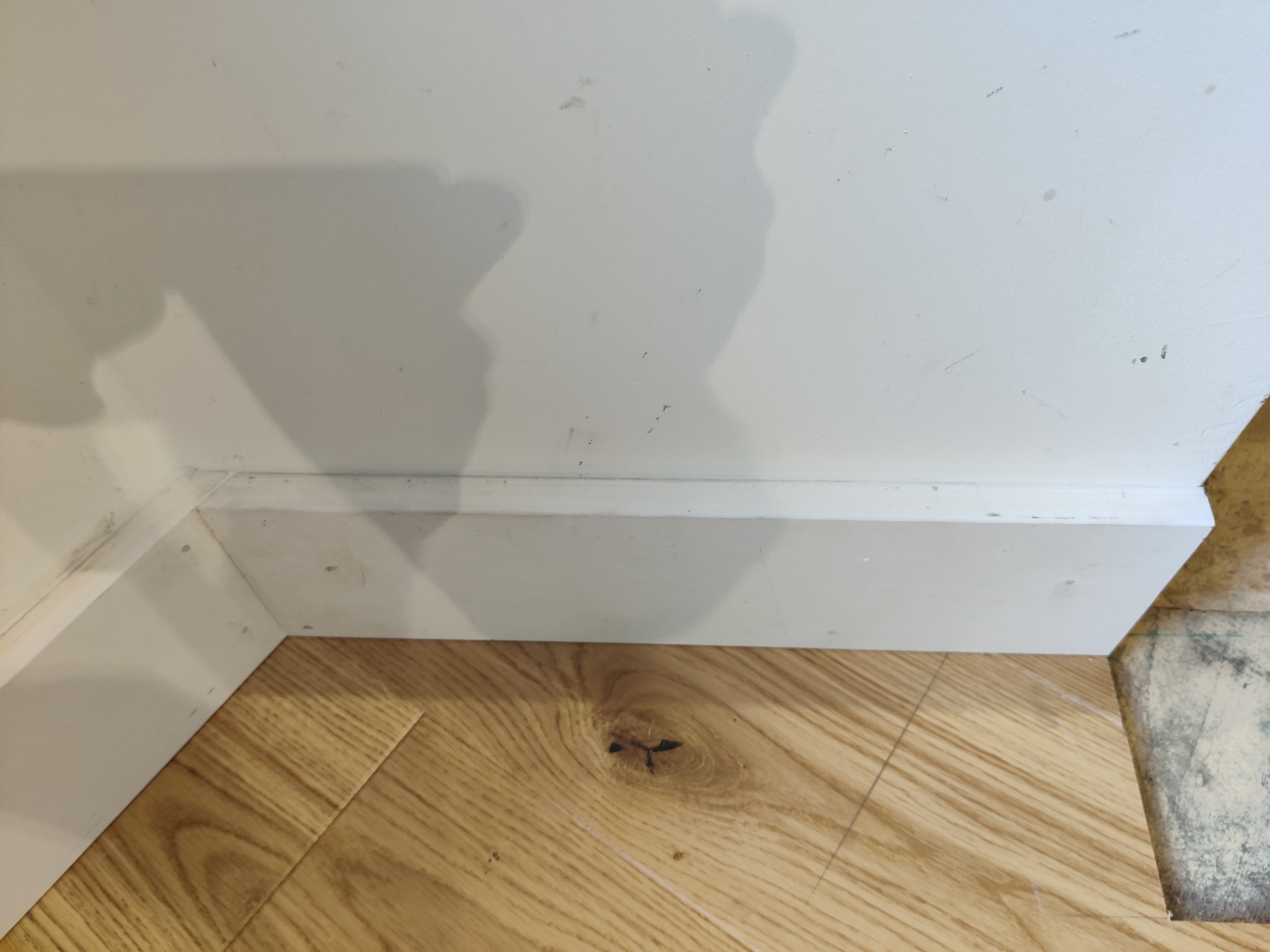 Fragment podłogi z jasnego drewna z widocznymi słojami i sękiem, biała listwa przypodłogowa przy brudnej, białej ścianie, widoczne niedociągnięcia w wykończeniu.