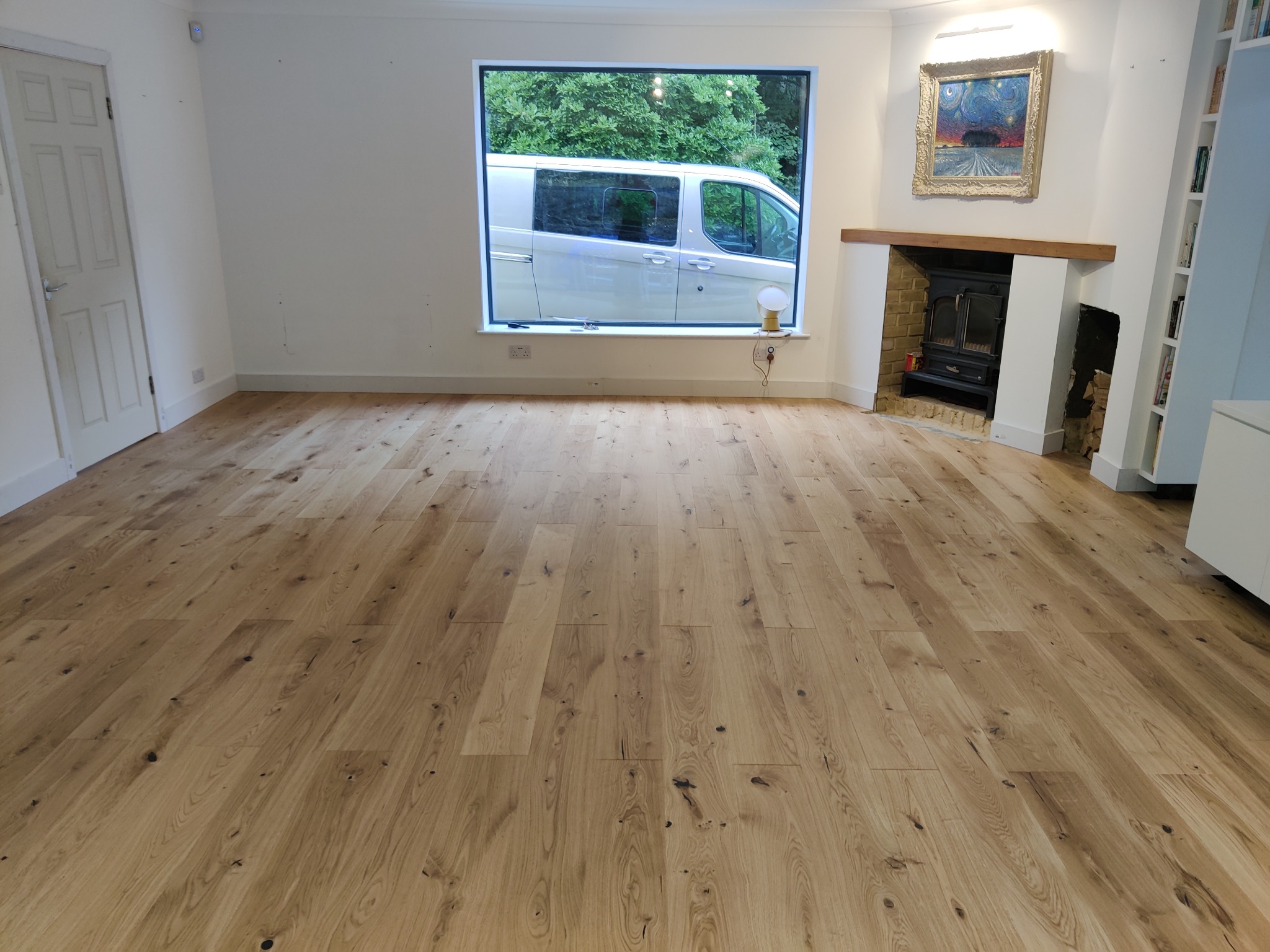 Jasny, drewniany parkiet ułożony w salonie z kominkiem i widokiem na samochód dostawczy za oknem.