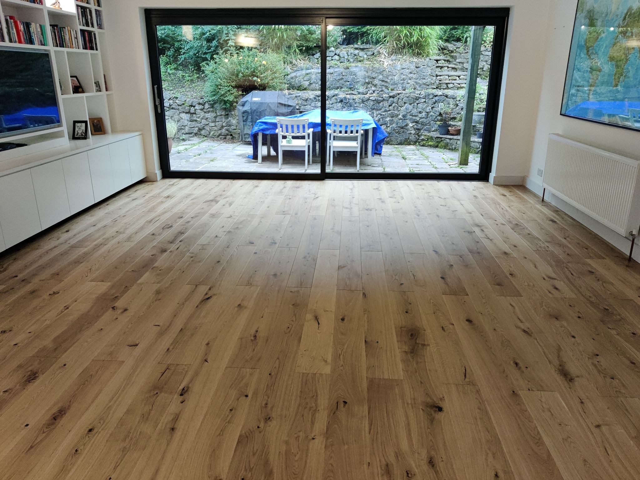 Jasna, nowa podłoga z desek parkietowych, widok na taras przez duże przesuwne okno, w tle kamienny murek i meble ogrodowe przykryte plandeką.