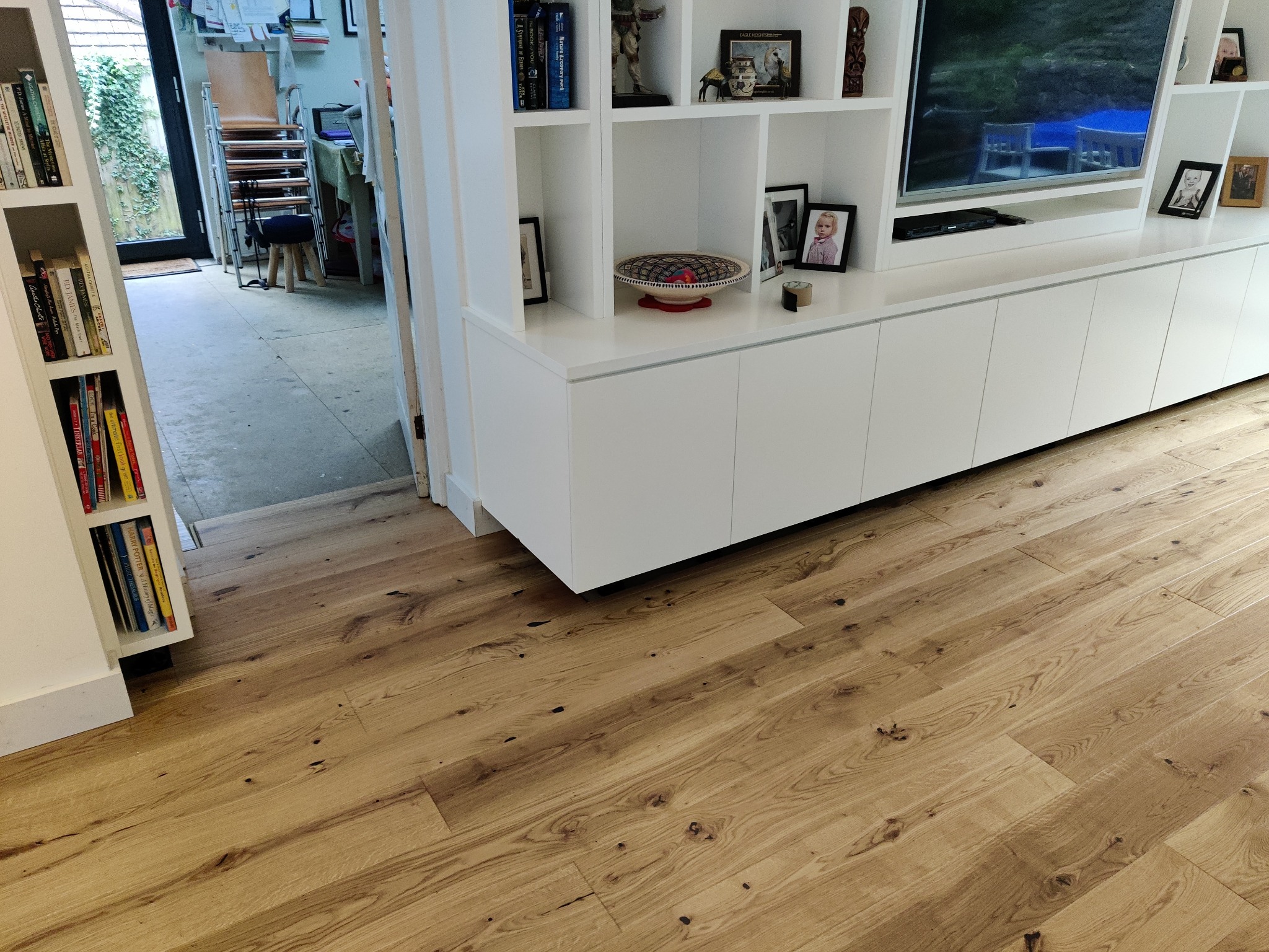 Jasny parkiet z widocznymi słojami drewna, ułożony w salonie z białymi meblami na wysoki połysk i regałem na książki, z widokiem na ogród przez otwarte drzwi.