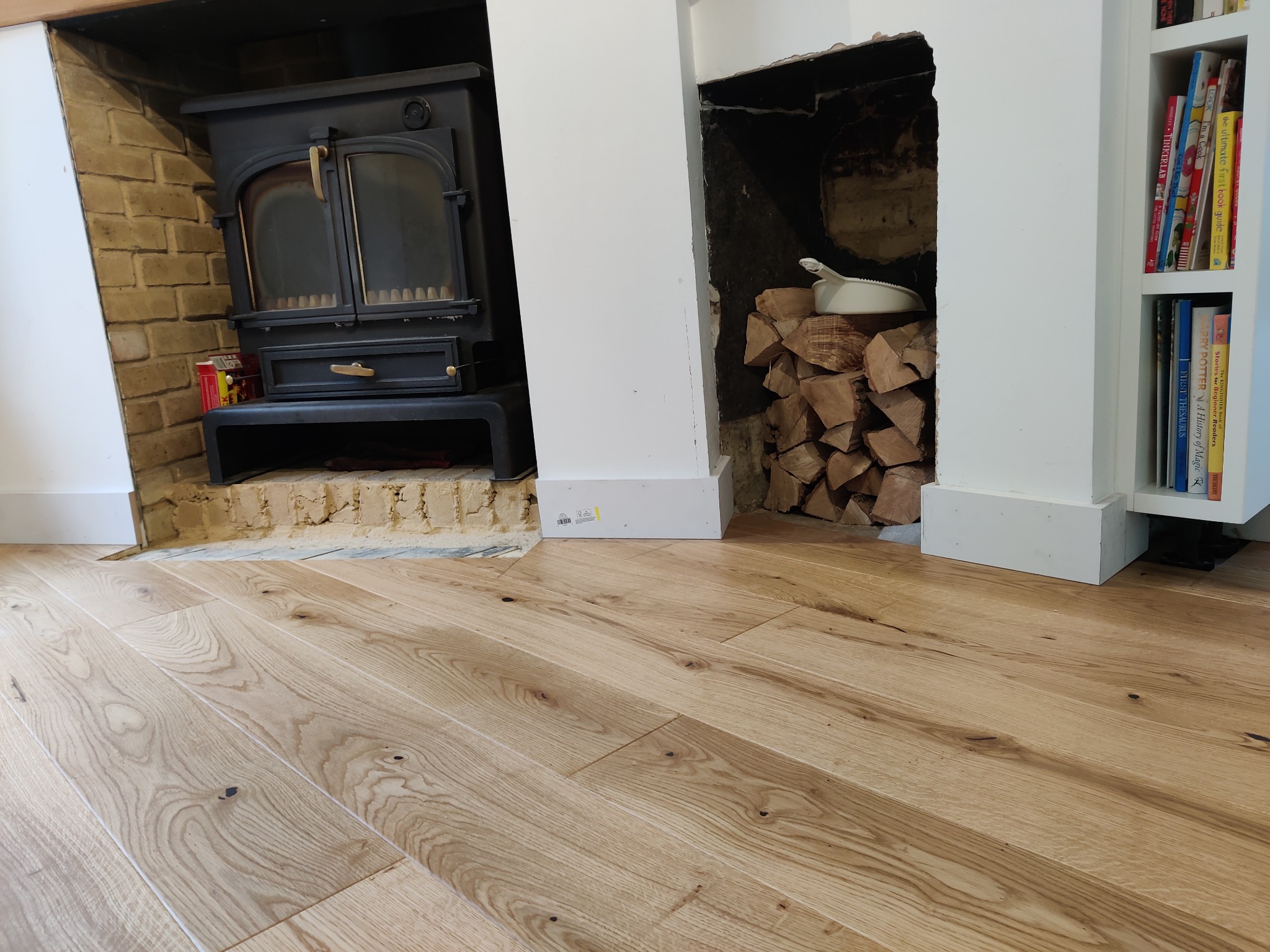 Jasne, drewniane panele podłogowe ułożone w salonie z kominkiem na drewno i półkami z książkami w tle.