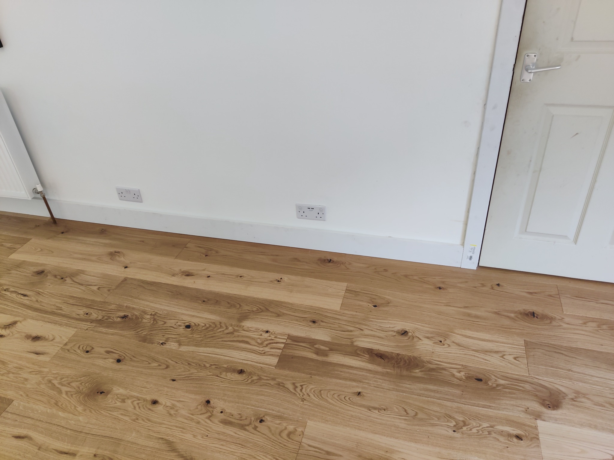Jasne panele podłogowe z widocznym rysunkiem drewna, biała ściana z dwoma gniazdkami elektrycznymi i fragment białych drzwi.