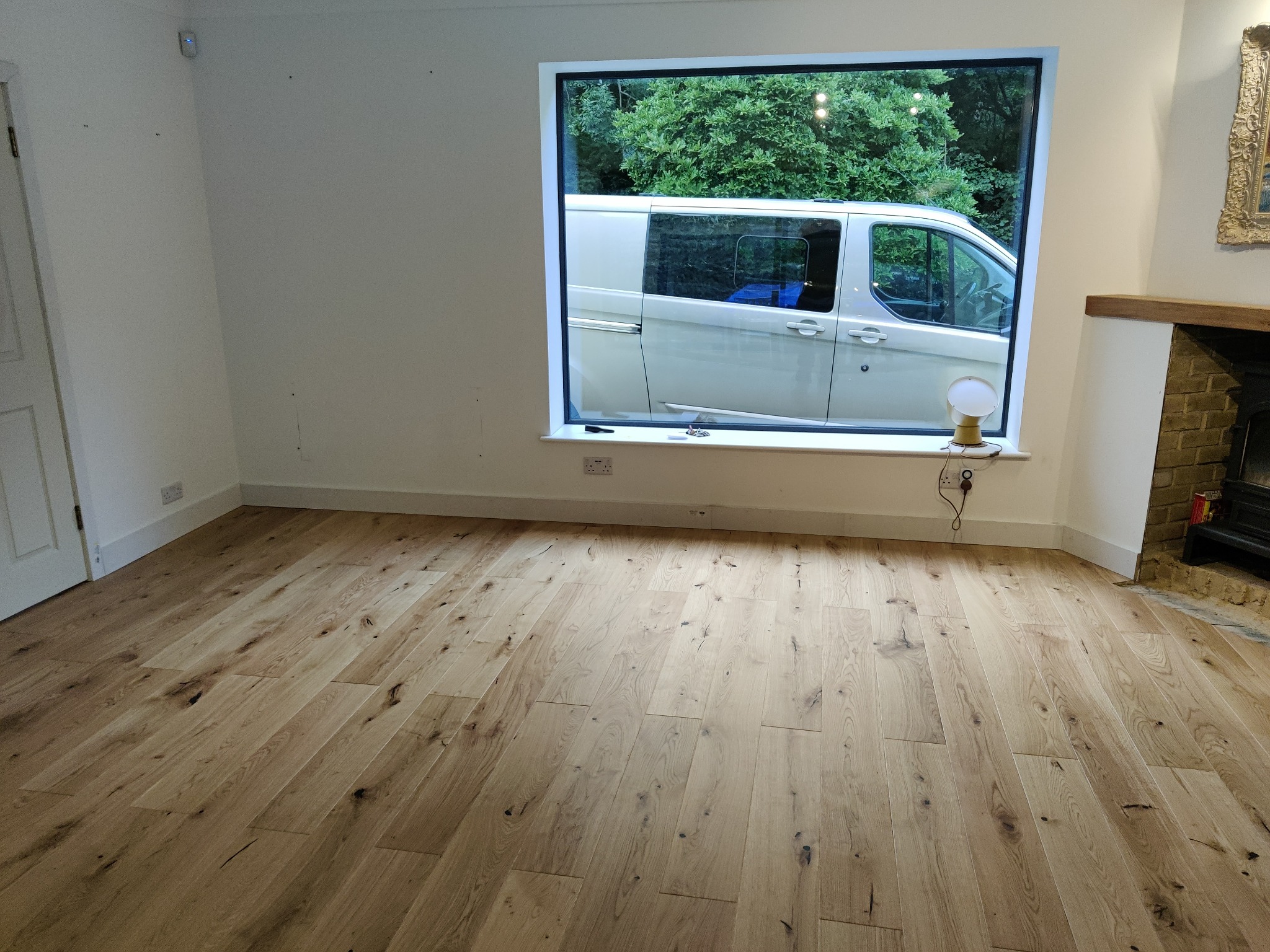 Jasne, naturalne drewniane deski podłogowe, ułożone ukośnie w przestronnym pokoju z białymi ścianami i widokiem na zieleń za oknem, z dostawczym samochodem zaparkowanym na zewnątrz.