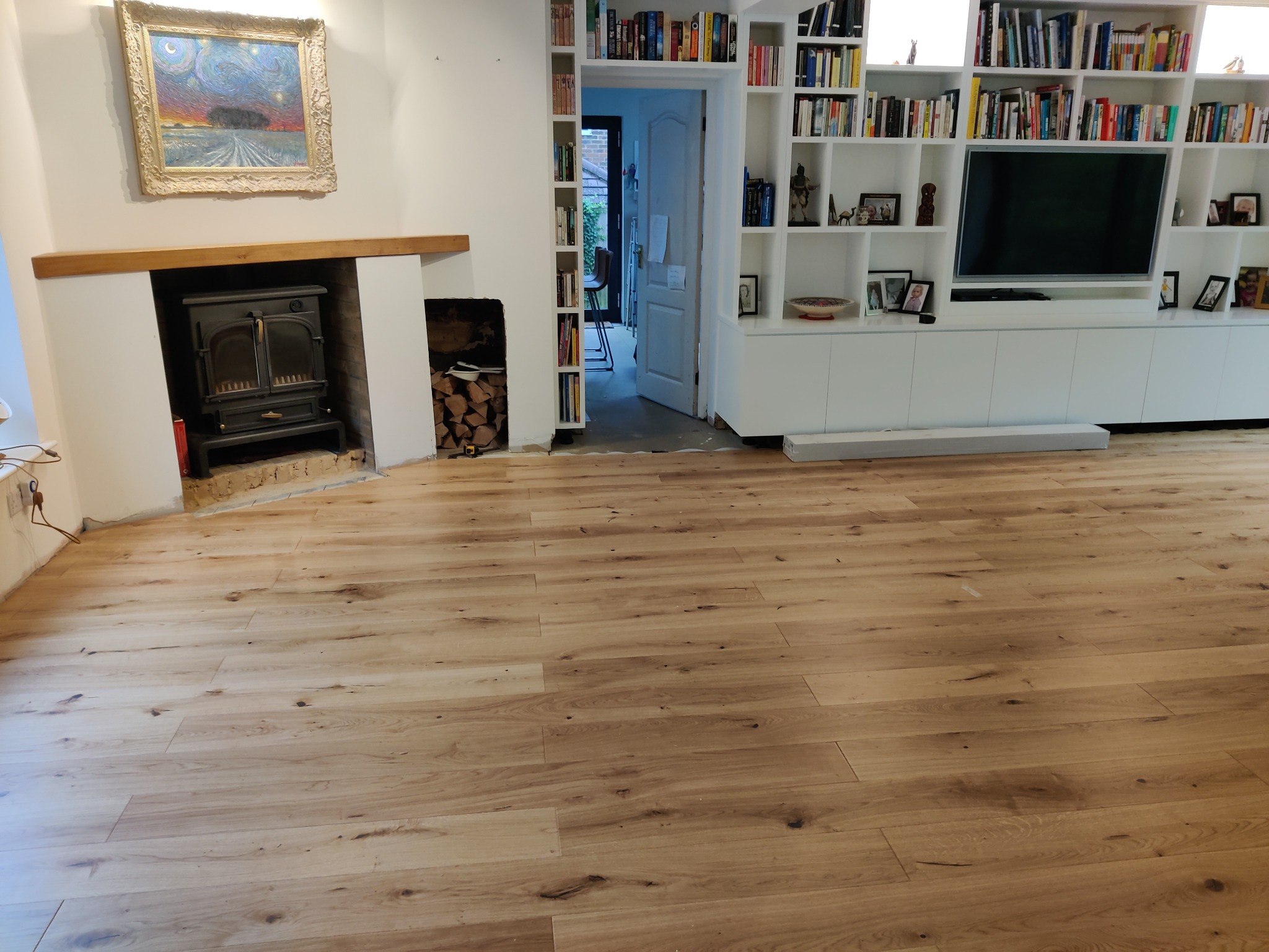 Jasne, drewniane deski podłogowe z wyraźnym usłojeniem, ułożone w salonie z kominkiem i regałem na książki, widoczne ślady po remoncie.