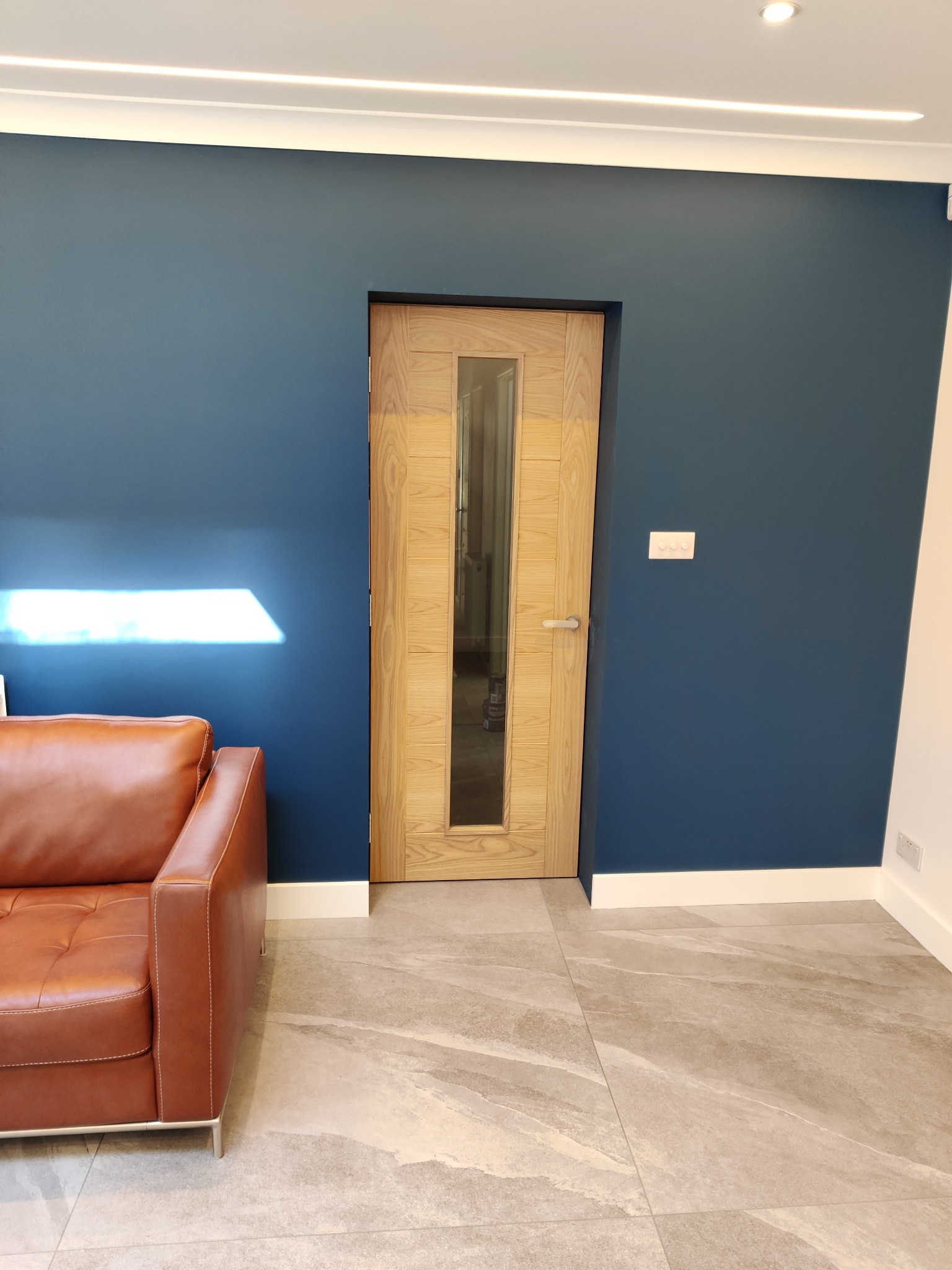 Wnętrze z ciemnoniebieską ścianą, drewnianymi drzwiami z szybą, fragmentem brązowej sofy i szarą podłogą z płytek imitujących kamień.