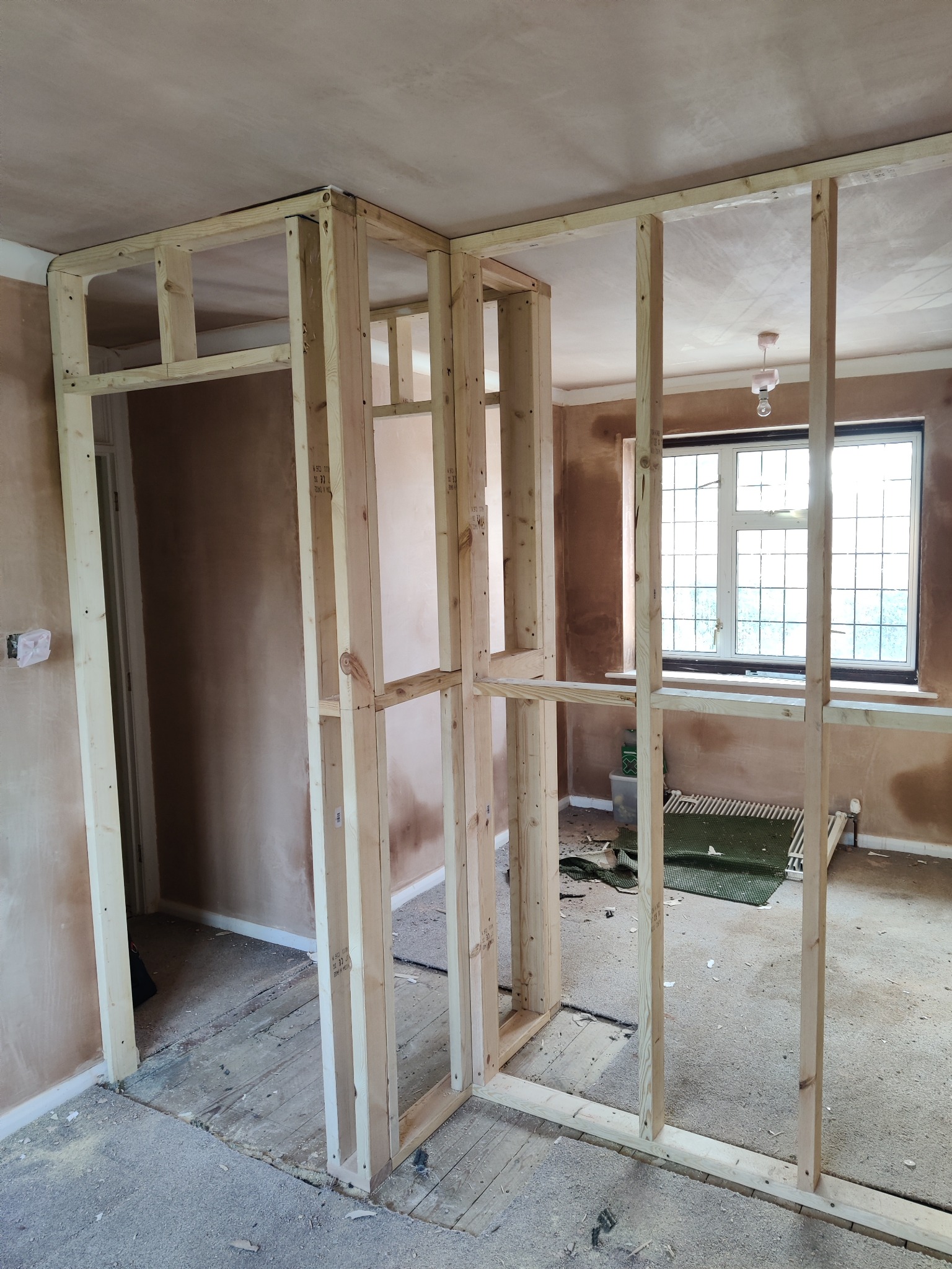 Wnętrze w trakcie remontu: drewniana konstrukcja szkieletowa ścianki działowej, widoczne surowe ściany i podłoga przed wykończeniem.