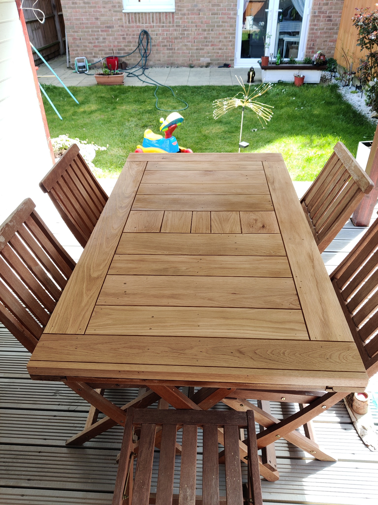 Drewniany stół ogrodowy z krzesłami na tarasie z desek kompozytowych, w tle trawnik i dom z cegły.