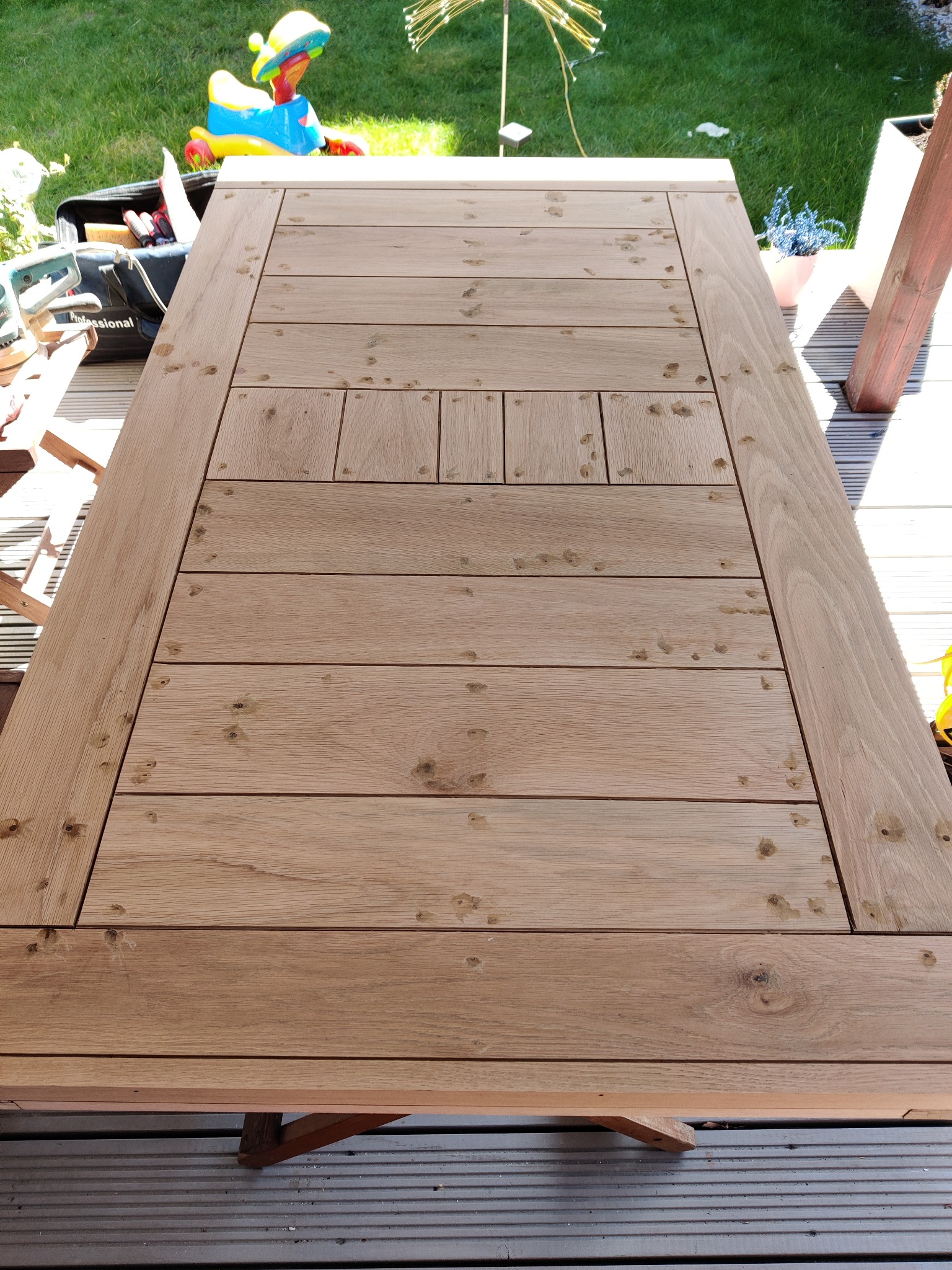 Detal drewnianego tarasu w trakcie budowy: widoczne deski z otworami po wkrętach, narzędzia w torbie obok, zabawka dziecięca w tle.