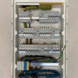 Rozdzielnica elektryczna z zamontowanymi bezpiecznikami i okablowaniem w białej obudowie, widoczne przewody w różnych kolorach i oznaczenia na listwach zaciskowych.