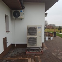 TechNovum Sp. z o.o. - Zewnętrzna jednostka klimatyzacji Panasonic Inverter zamontowana na elewacji domu z brązowym dachem, widoczna mniejsza jednostka Midea, na pierwszym planie kostka brukowa.