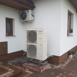 TechNovum Sp. z o.o. - Zewnętrzna jednostka klimatyzacji Panasonic Inverter, dwuczęściowa, zamontowana na metalowym stelażu na posesji z kostki brukowej, z widoczną jednostką wewnętrzną Midea na ścianie budynku z oknami.