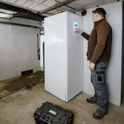 TechNovum Sp. z o.o. - Instalator z czołówką dotyka ekranu białej pompy ciepła w piwnicy, obok leży czarna skrzynka narzędziowa.