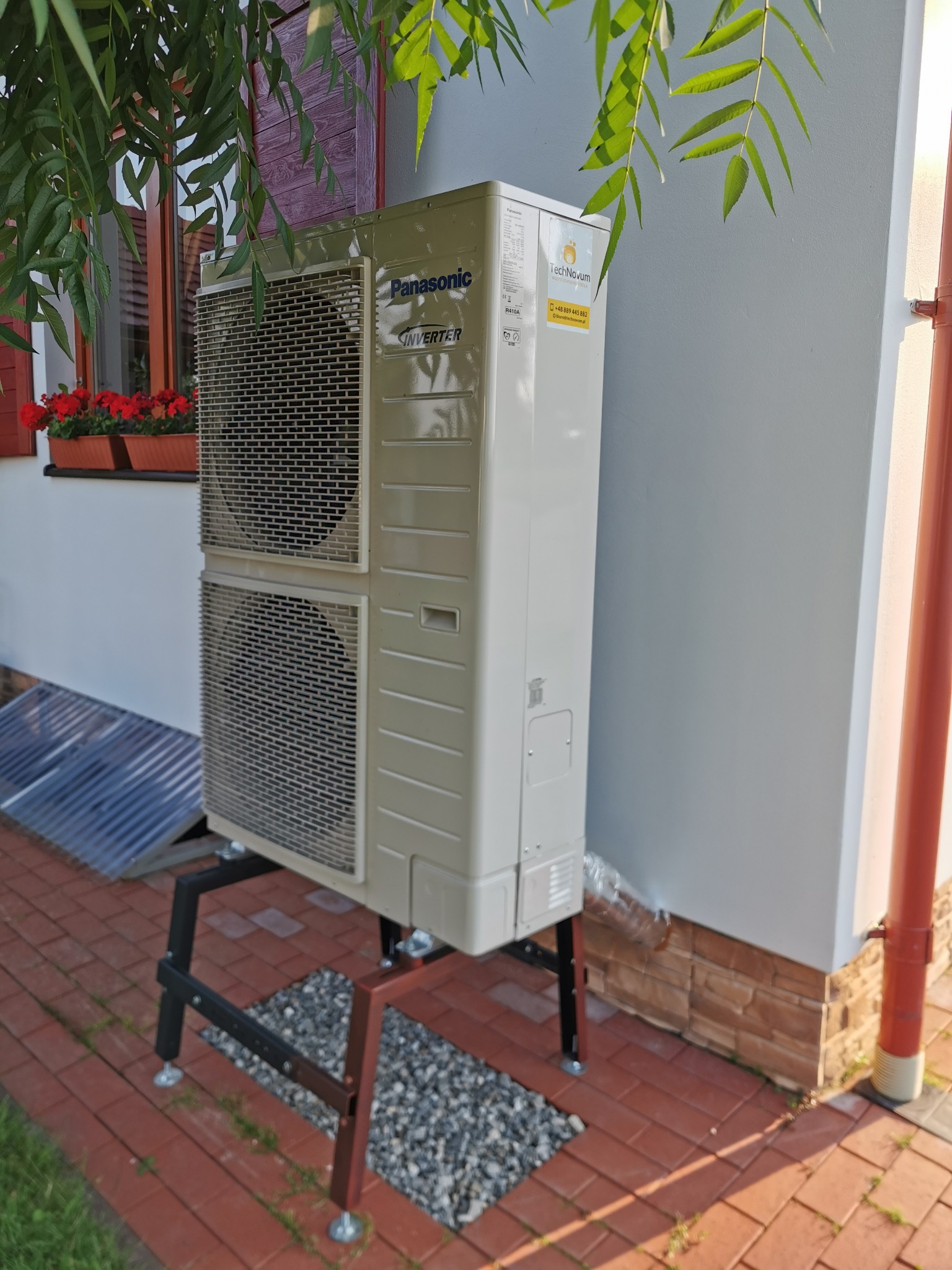Zewnętrzna jednostka klimatyzacji Panasonic Inverter na metalowym stelażu, widoczna na tle białej ściany domu z czerwonymi okiennicami i kwiatami w doniczkach.