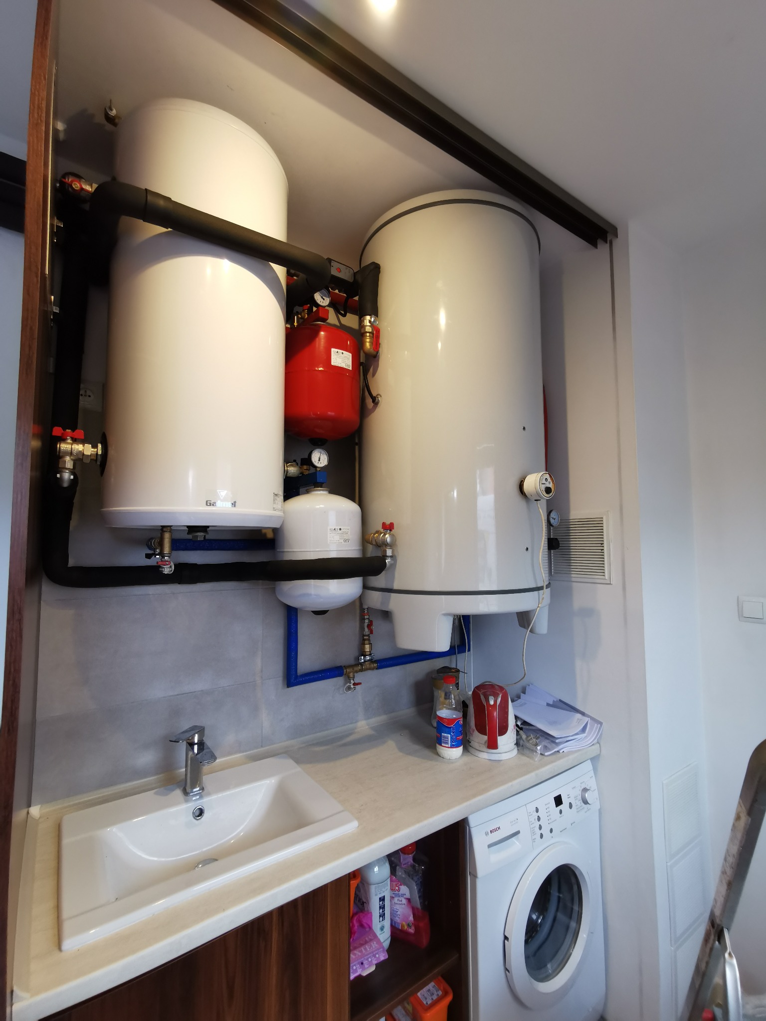 Kompaktowa instalacja zbiorników buforowych i zasobnika ciepłej wody z widocznymi rurami w izolacji, zamontowana w pomieszczeniu gospodarczym nad pralką i zlewem.