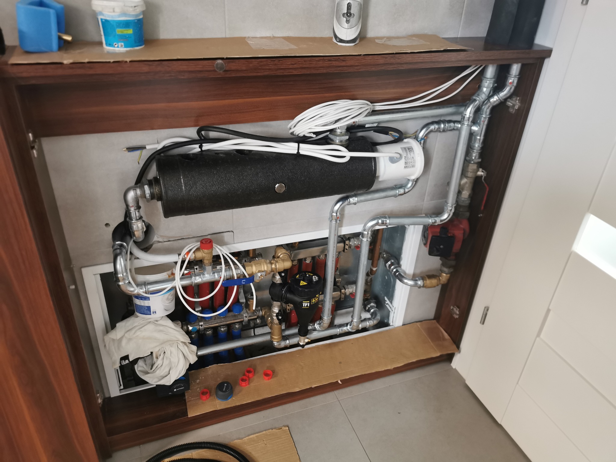 Rozdzielacz ogrzewania podłogowego w szafce z brązowego drewna, widoczne liczne rury, zawory, czujnik TPI, białe kable i pojemnik z farbą na górze.