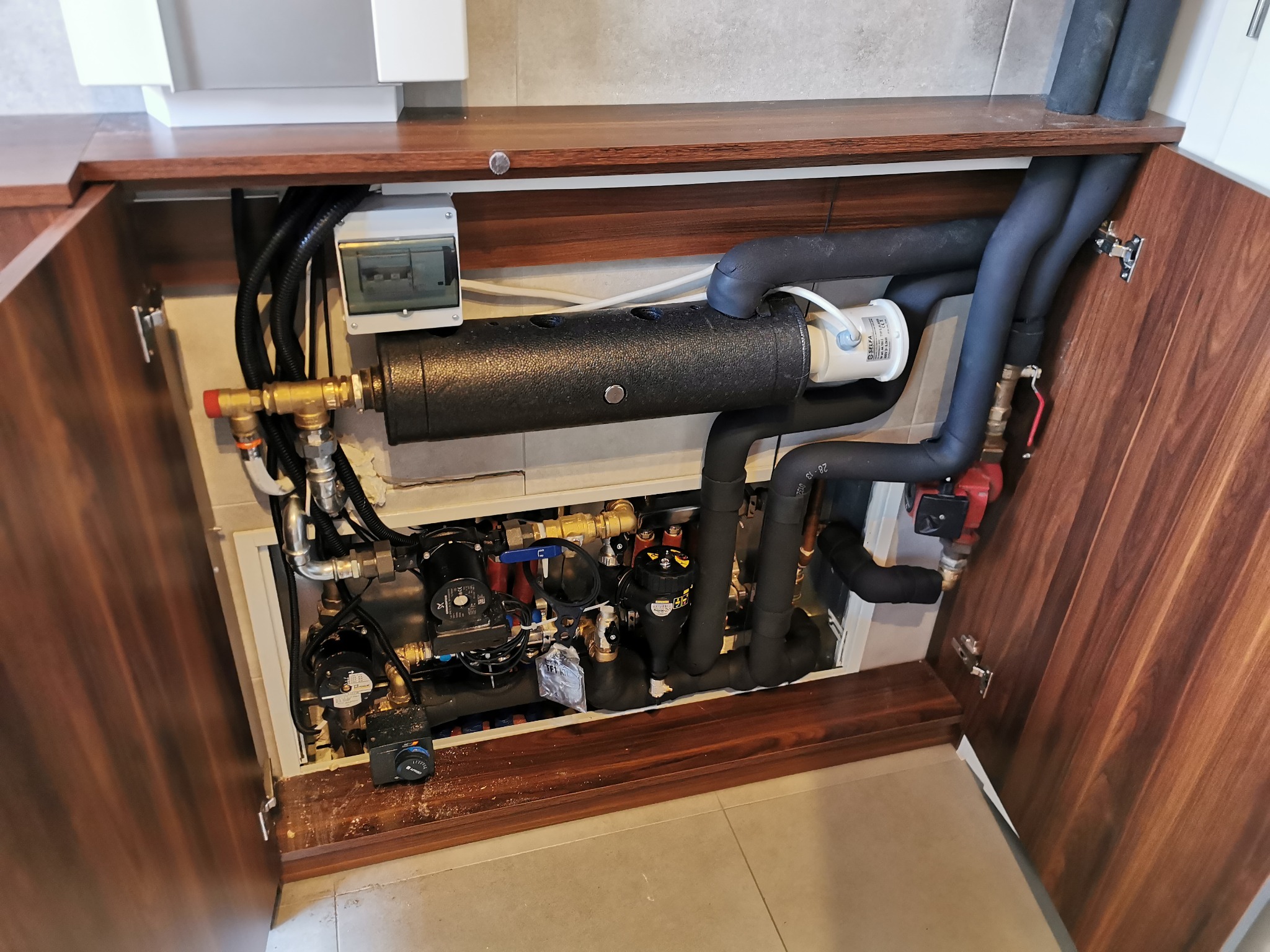 Węzeł cieplny z czarnymi rurami izolowanymi termicznie, pompami obiegowymi i osprzętem schowany w szafce z drewnianymi frontami.