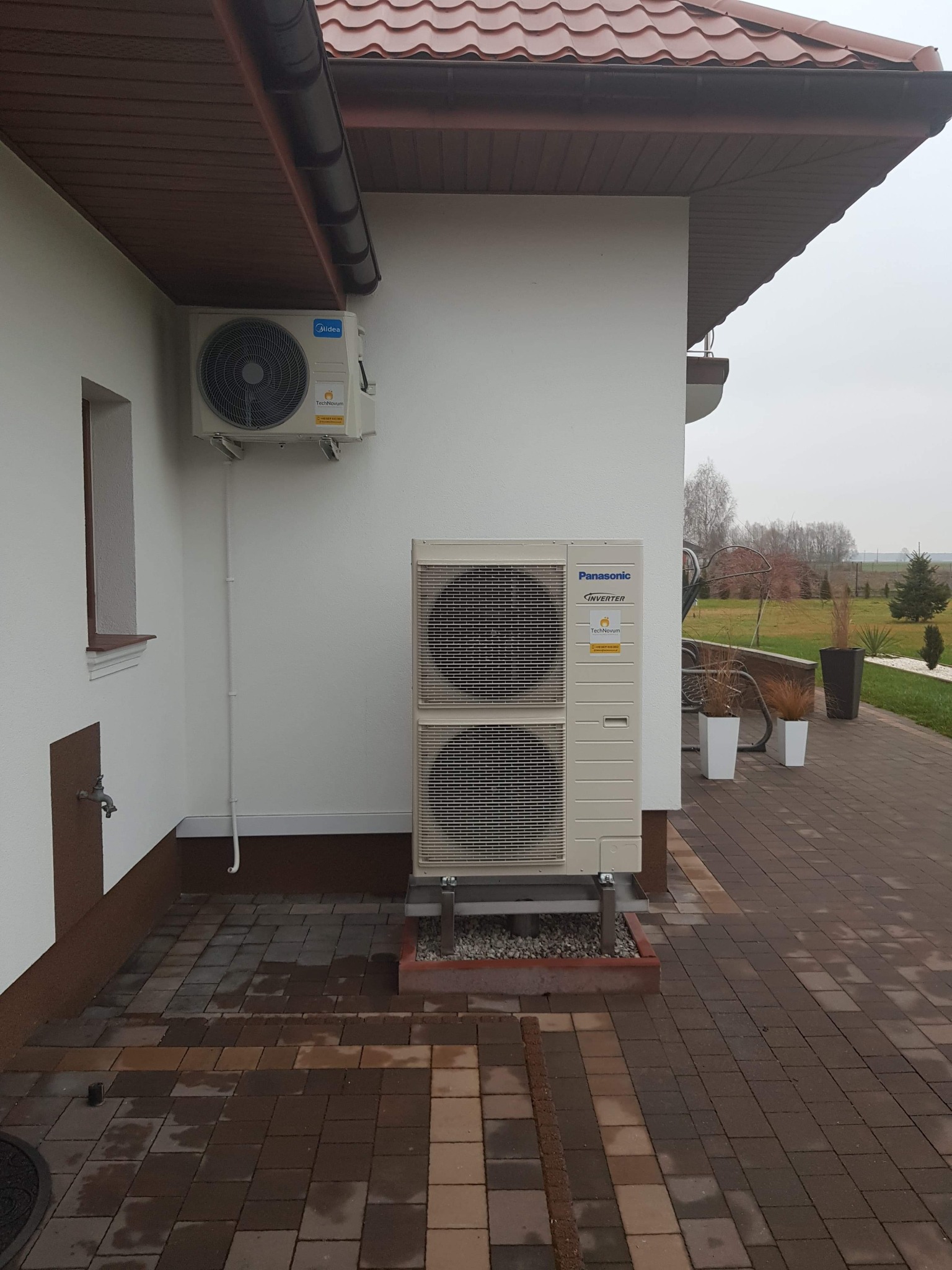 Zewnętrzna jednostka klimatyzacji Panasonic Inverter zamontowana na elewacji domu z brązowym dachem, widoczna mniejsza jednostka Midea, na pierwszym planie kostka brukowa.