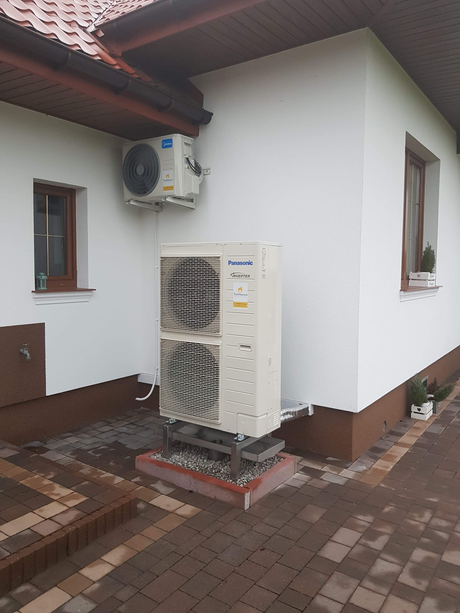 Zewnętrzna jednostka klimatyzacji Panasonic Inverter, dwuczęściowa, zamontowana na metalowym stelażu na posesji z kostki brukowej, z widoczną jednostką wewnętrzną Midea na ścianie budynku z oknami.