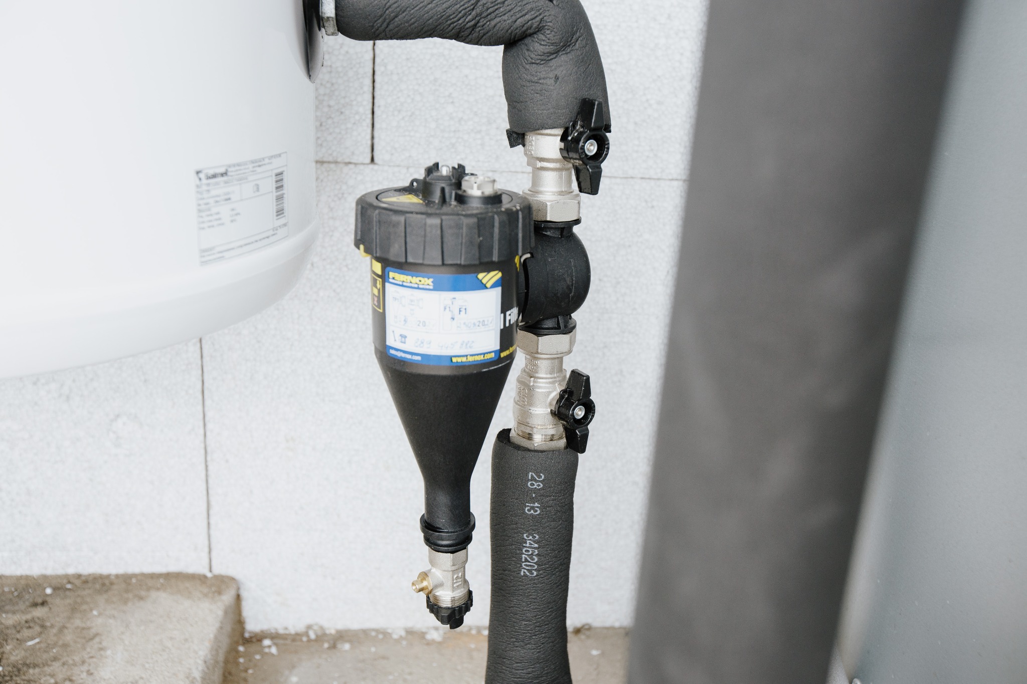 Filtr magnetyczny Fernox TF1 zamontowany na rurach instalacji grzewczej, z widocznymi zaworami i izolacją termiczną, na tle ściany z bloczków betonowych.