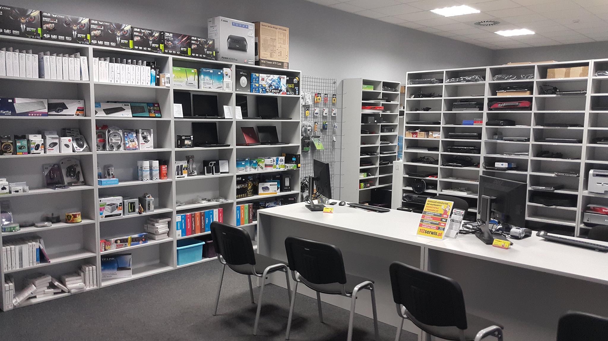 Wnętrze sklepu komputerowego z białymi regałami wypełnionymi sprzętem i akcesoriami, za ladą trzy krzesła dla klientów i plakat z napisem 'Oddałeś komputer do naprawy? Skorzystaj z promocji'.