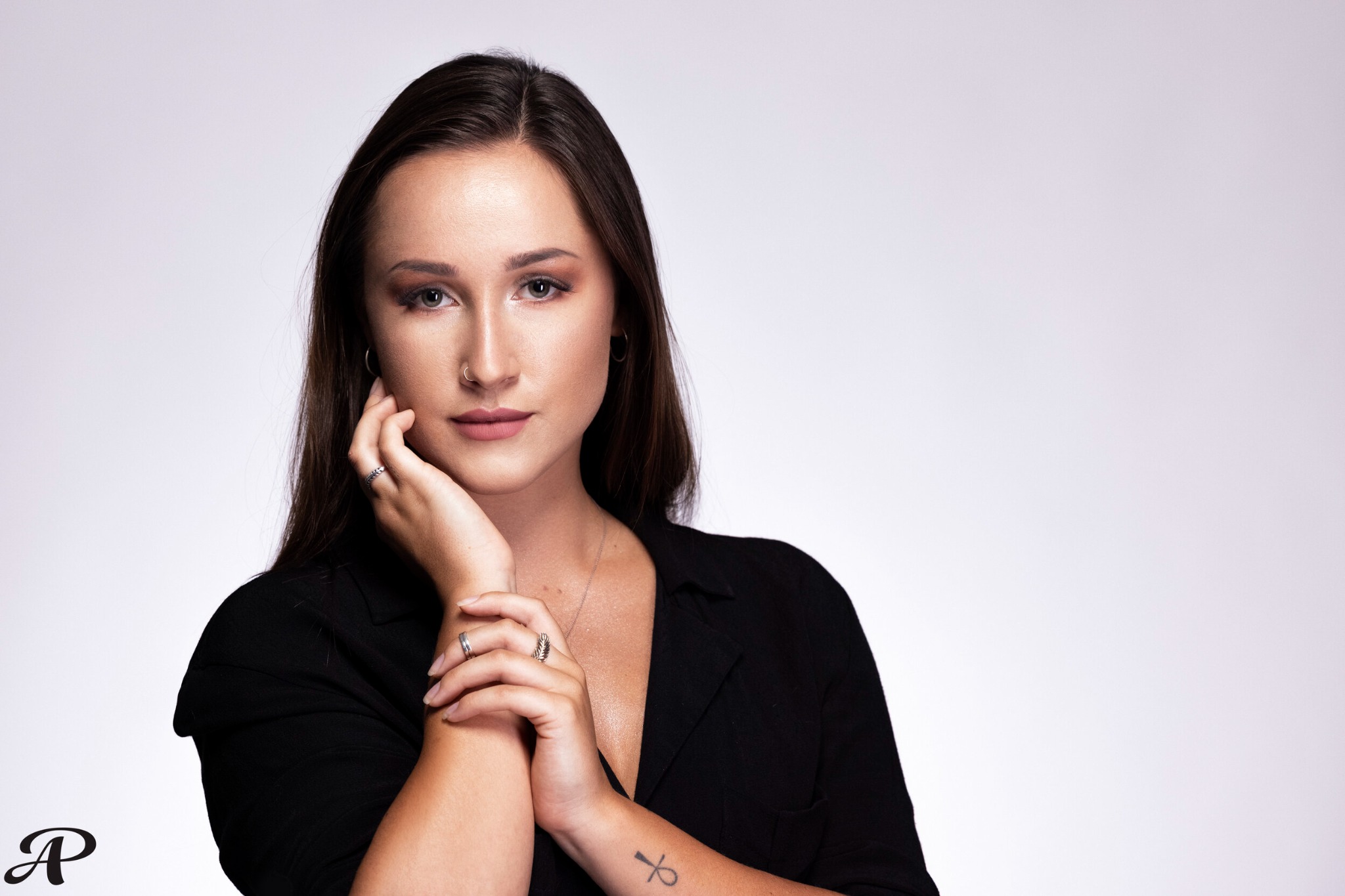 Portret kobiety z nosem przekłutym kolczykiem, dłonie w pobliżu twarzy, tatuaż ankh na przedramieniu, pierścionki na palcach, czarna koszula na jasnym tle.