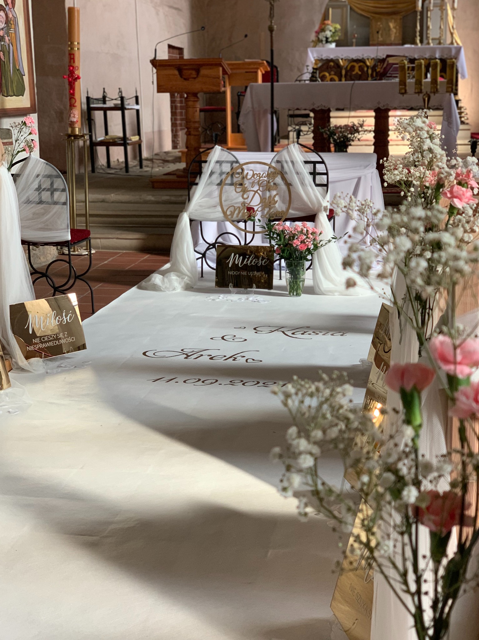 Dekoracja kościoła na uroczystość ślubną: biały dywan z napisem imion nowożeńców i datą, ozdobne tabliczki z cytatami o miłości, kompozycje kwiatowe w odcieniach różu i bieli, krzesła udekorowane...