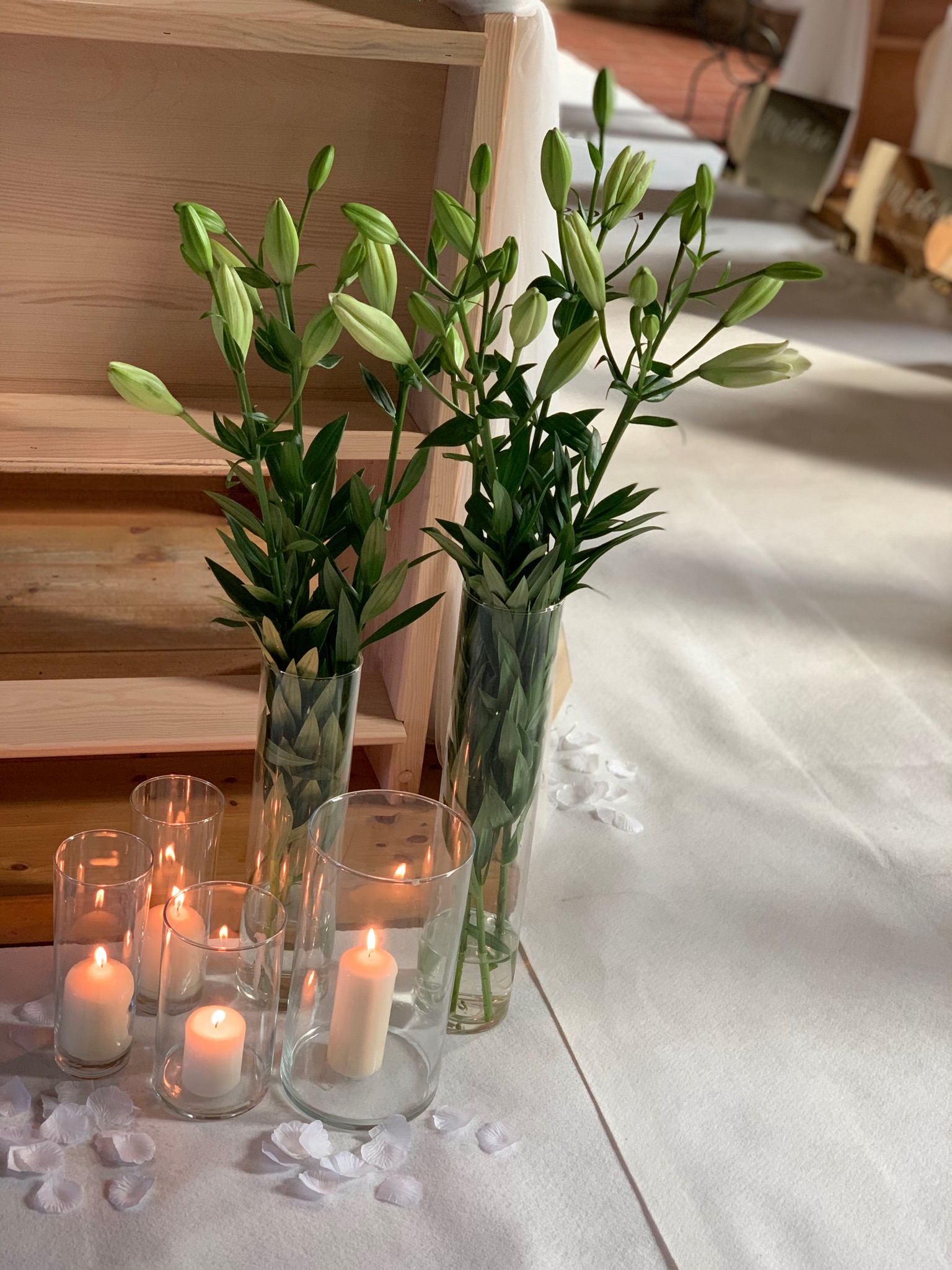 Dekoracja stołu z wysokimi wazonami z liliami w pąkach i świecami w szklanych osłonkach, rozrzucone białe płatki.