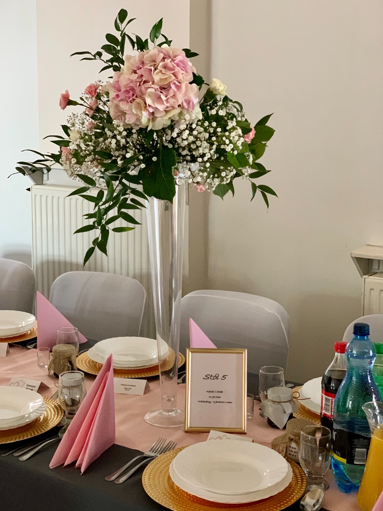 Dekoracja stołu weselnego: wysoki szklany wazon z kompozycją kwiatową z hortensji i gipsówki, złote podkładki pod talerze, różowe serwetki, woda w butelkach i ramka ze spersonalizowanym napisem...