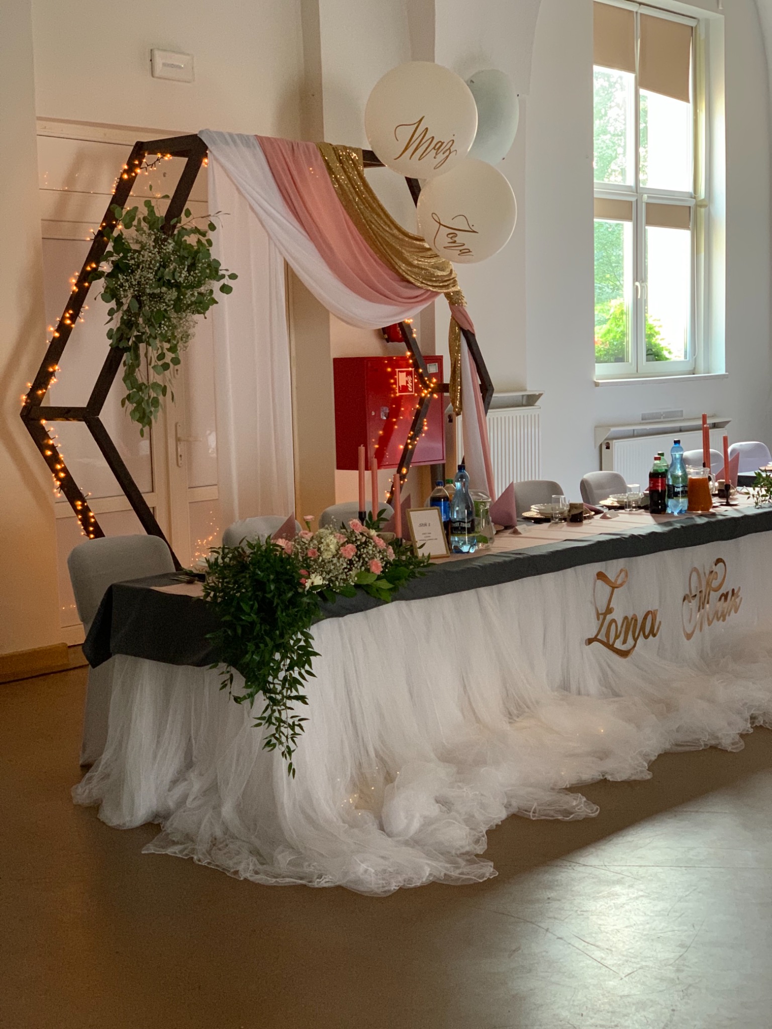 Udekorowany stół weselny z czarnym blatem, białym tiulowym obrusem, złotymi napisami 'Żona Mąż', kwiatami, świecami i dekoracją z drewnianej konstrukcji z lampkami, tkaninami i balonami z napisami.
