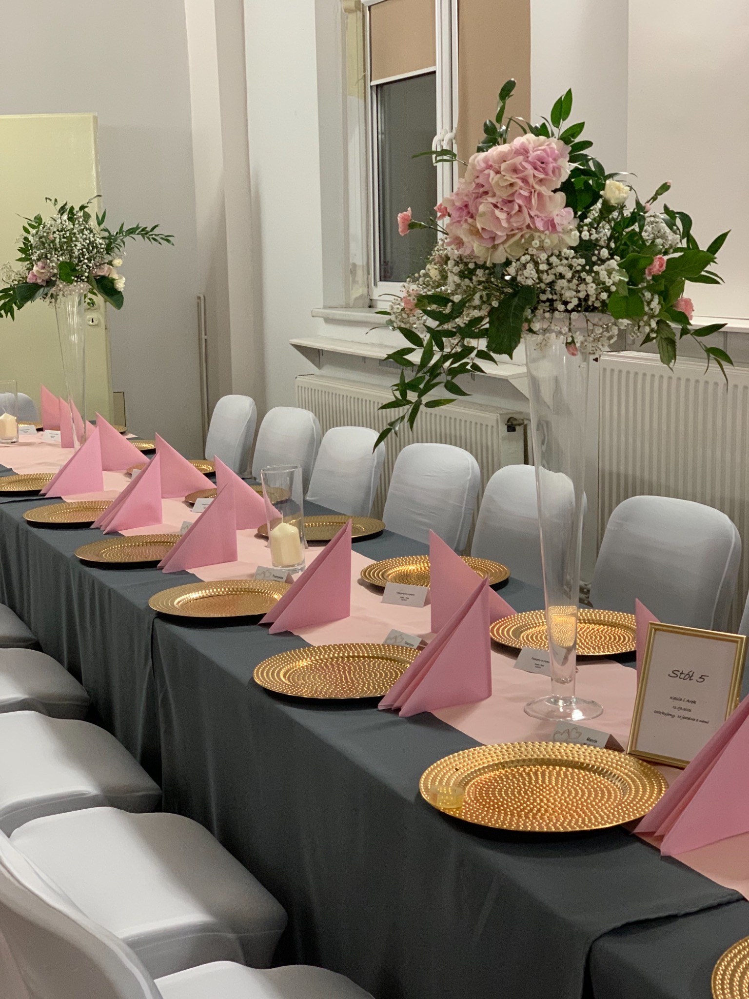 Dekoracja stołu weselnego z różowymi serwetkami, złotymi talerzami i wysokimi wazonami z kwiatami, ustawiona w sali z białymi krzesłami.