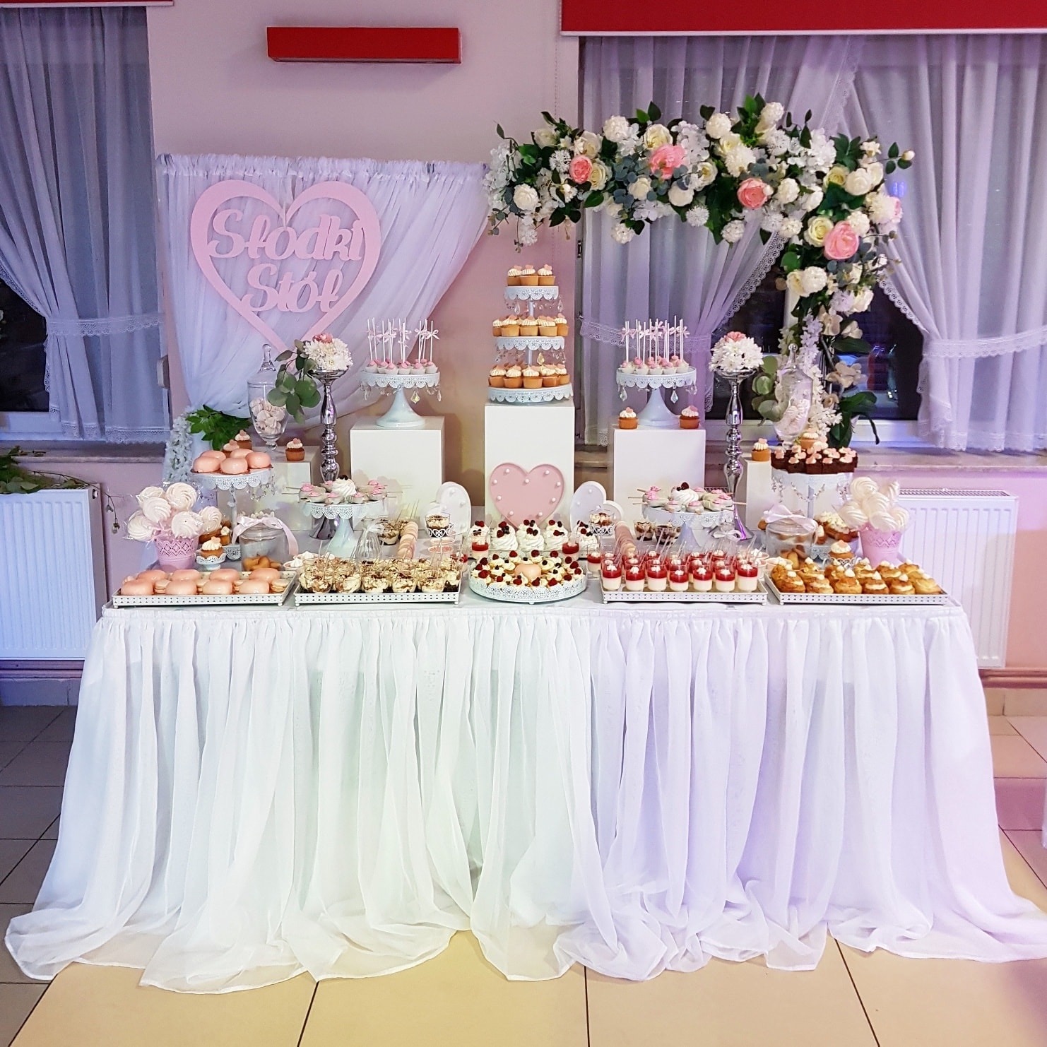 Bogato zastawiony stół z różnorodnymi słodkościami, w tym babeczki, desery w szklankach i lizaki, udekorowany białym obrusem i kwiatową girlandą, z dekoracyjnym sercem z napisem 'Słodki Stół' w tle.