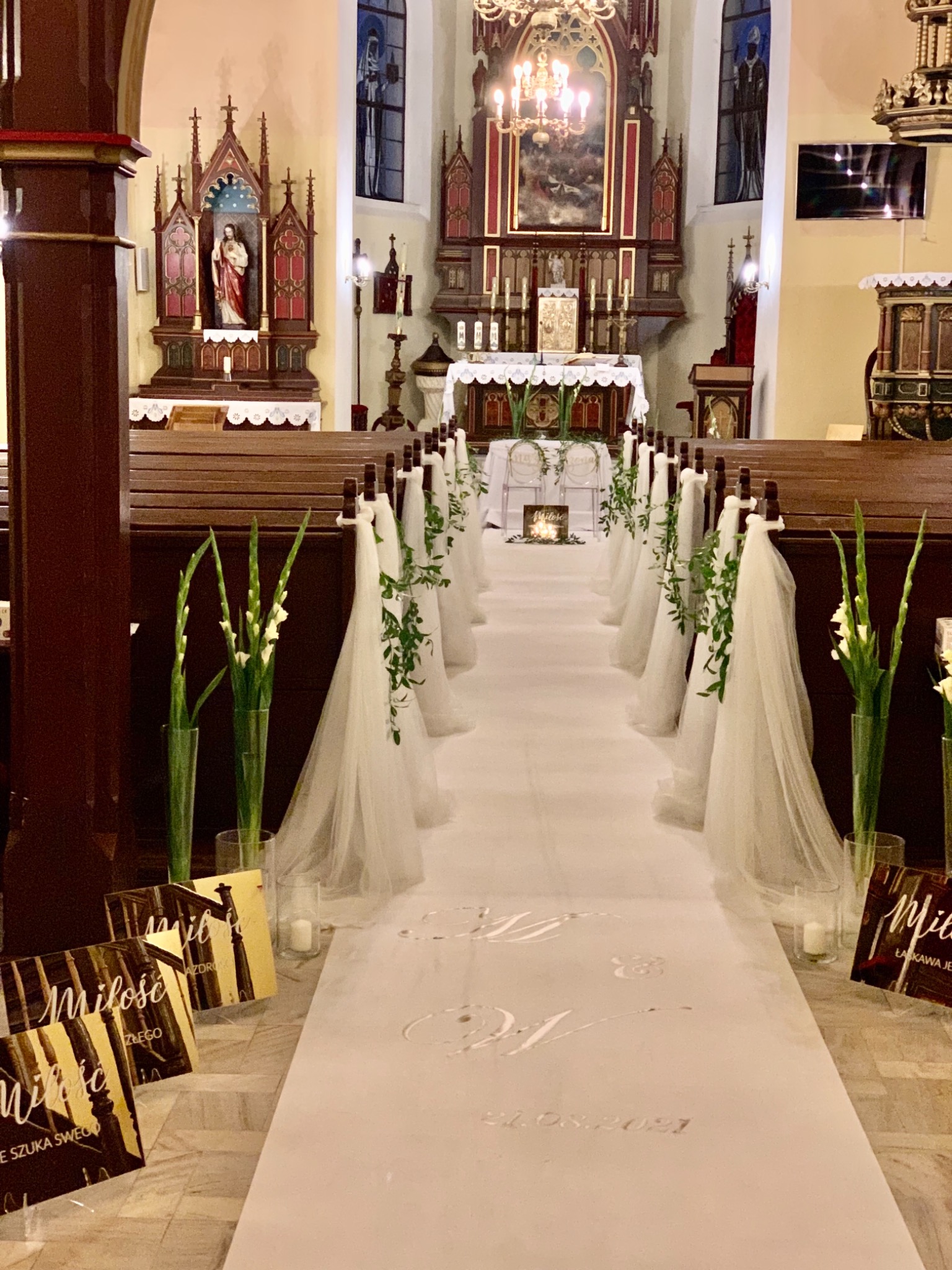 Kościół udekorowany na ślub: biały dywan z napisem 'Miłość' i datą, białe tiule i kwiaty zdobią ławki, w tle ołtarz
