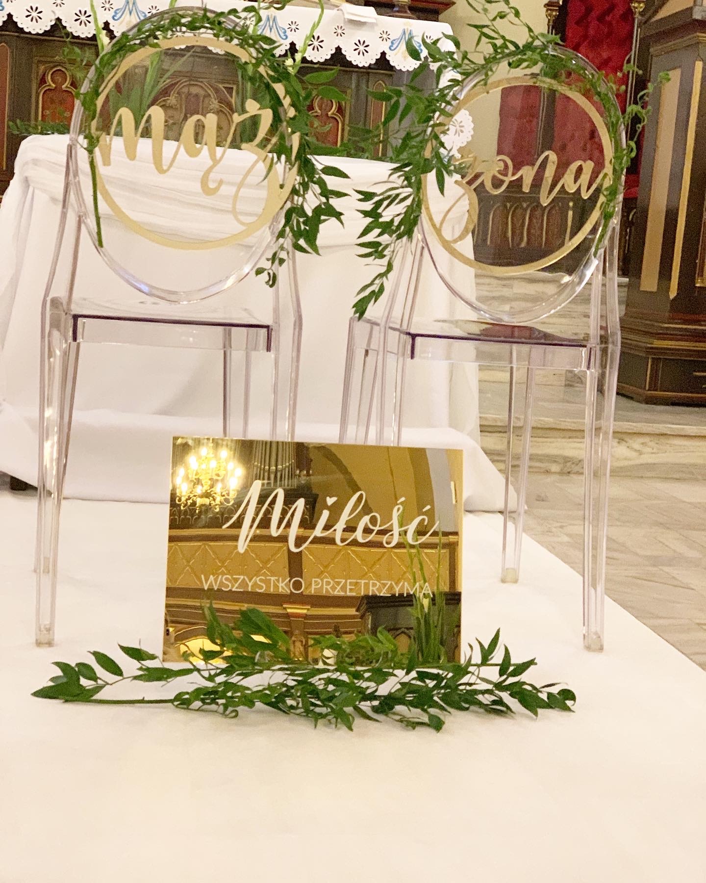 Transparentne krzesła z dekoracjami ślubnymi: okrągłe napisy 'Mąż' i 'Żona' otoczone zielonymi gałązkami, złoty napis 'Miłość wszystko przetrzyma' odbijający żyrandol.