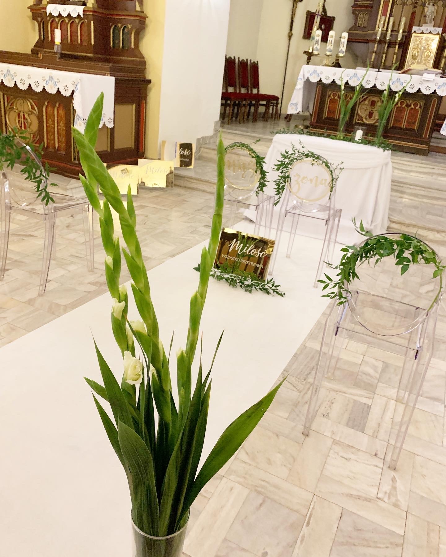 Dekoracja kościoła na uroczystość ślubną: bukiet mieczyków w szklanym wazonie na białym dywanie, przezroczyste krzesła z zielonymi girlandami i złotymi napisami 'Mąż' i 'Żona', w tle ołtarz.