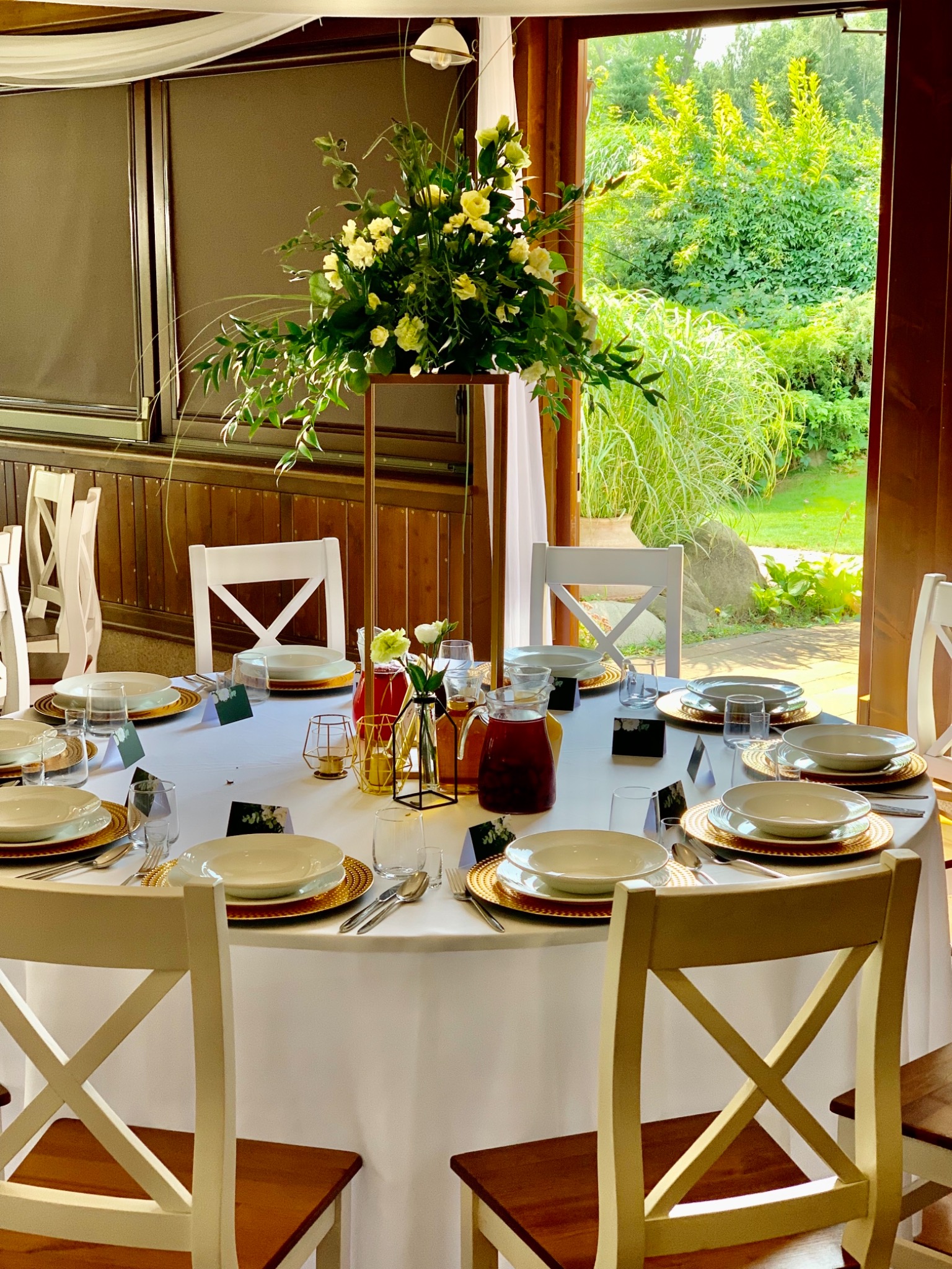 Elegancko nakryty okrągły stół weselny z białym obrusem, złotymi podkładkami, białymi talerzami, szklankami i sztućcami, wysoka kompozycja kwiatowa w złotym stojaku, białe krzesła z drewnianymi...