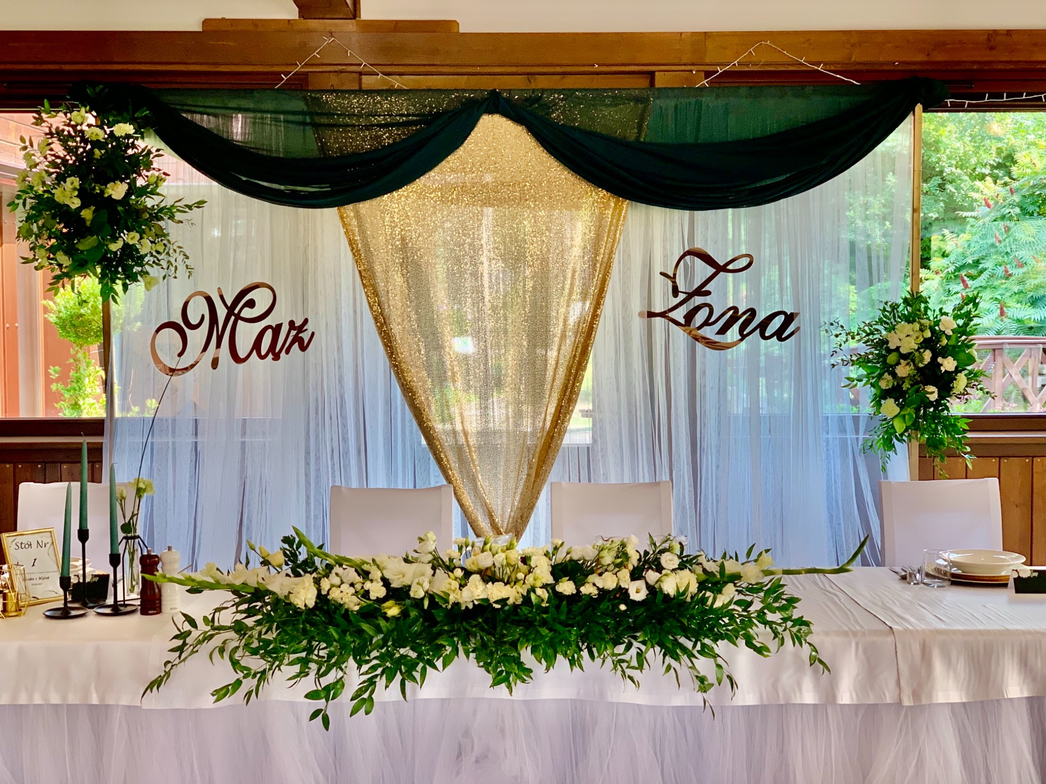 Dekoracja stołu weselnego z białymi kwiatami, złotym, błyszczącym tłem i zielonymi drapowanymi tkaninami, z napisem 'Mąż' i 'Żona' w tle.