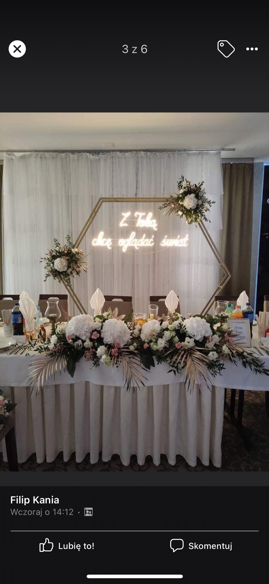 Dekoracja stołu weselnego z białymi kwiatami, złotymi liśćmi i neonowym napisem w geometrycznej ramie, na tle białej zasłony.