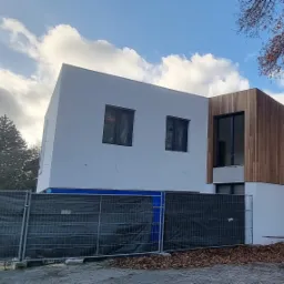 Nowoczesny dom dwukondygnacyjny z białą elewacją i drewnianą okładziną, zabezpieczony siatką budowlaną, widok z zewnątrz w pochmurny dzień.