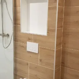 Nowoczesna łazienka z wiszącą toaletą, drewnopodobnymi płytkami i wnęką w ścianie, widoczny fragment kabiny prysznicowej.