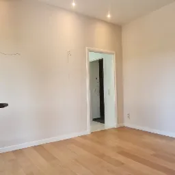 Pusty pokój z drewnianą podłogą, świeżo pomalowanymi ścianami w odcieniach beżu i dwiema lampami sufitowymi, widok na otwarte drzwi do kolejnego pomieszczenia.