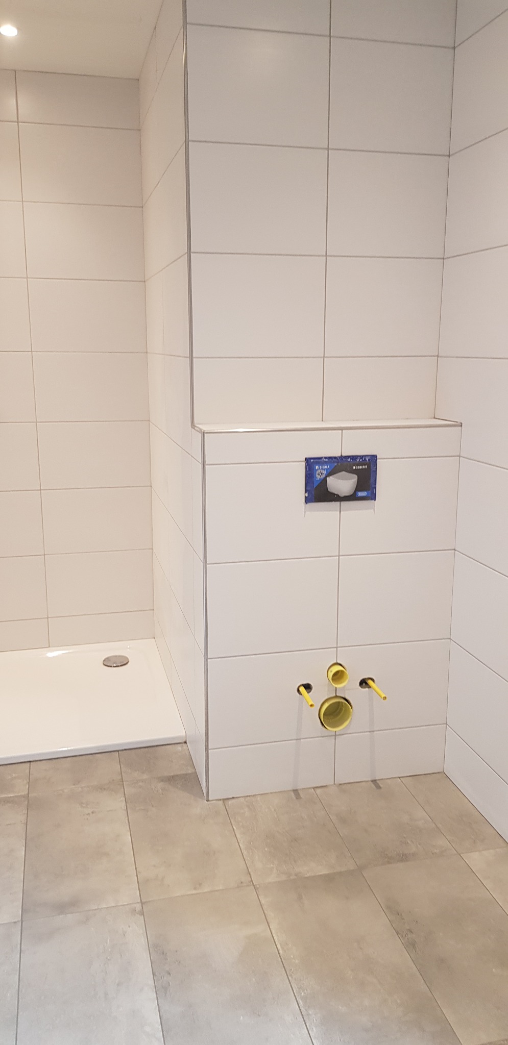 Fragment łazienki w trakcie remontu: białe płytki ścienne z szarymi fugami, zamontowany odpływ prysznicowy i żółte rury kanalizacyjne, szara podłoga z płytek imitujących kamień.