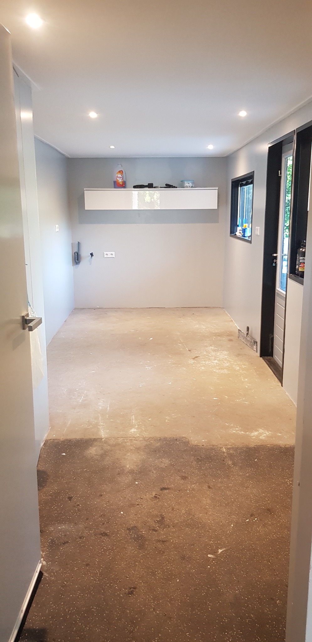 Wnętrze pokoju w trakcie remontu: szare ściany, betonowa podłoga w dwóch odcieniach, biała szafka wisząca i czarna rama okienna.