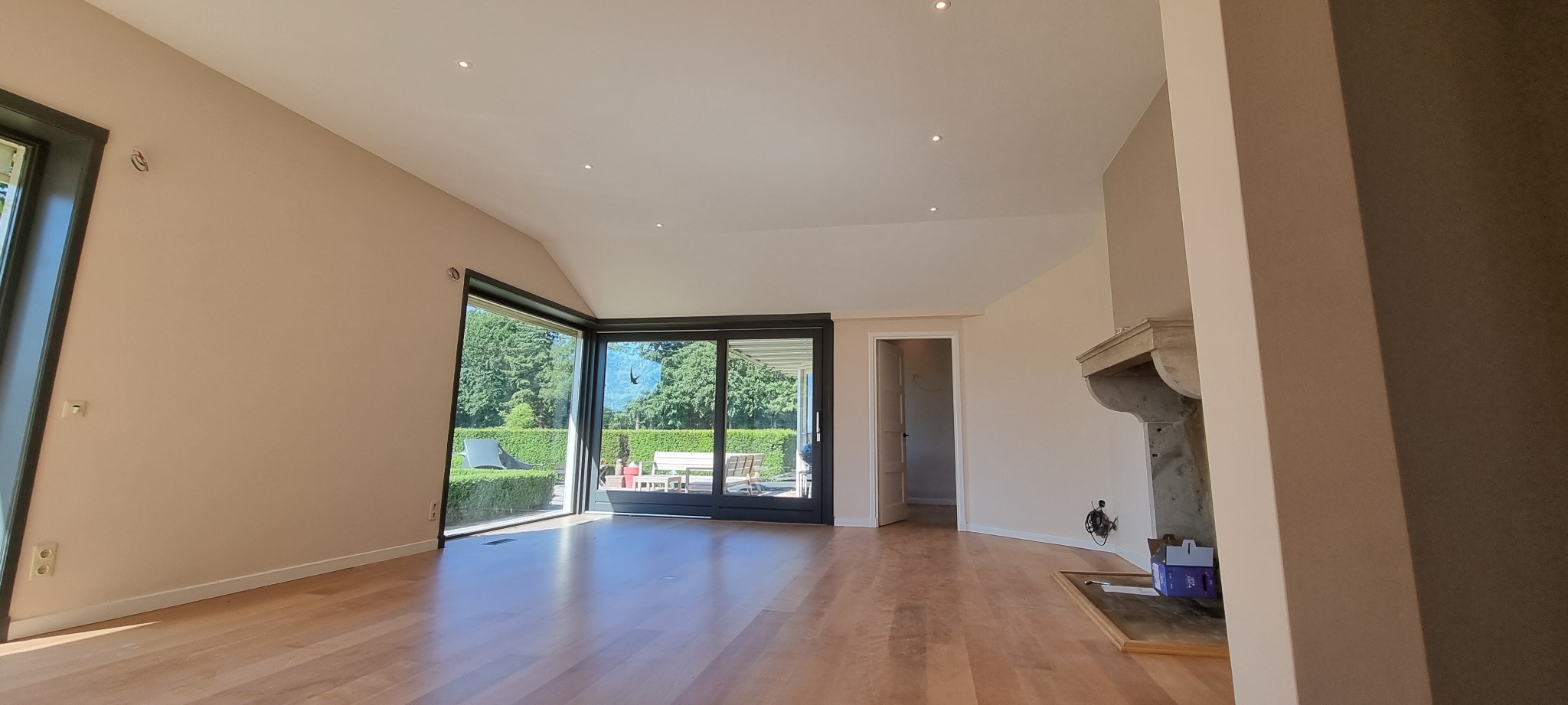 Jasne, przestronne wnętrze z drewnianą podłogą, dużymi przesuwnymi oknami z widokiem na ogród, kremowymi ścianami i ozdobnym kominkiem.